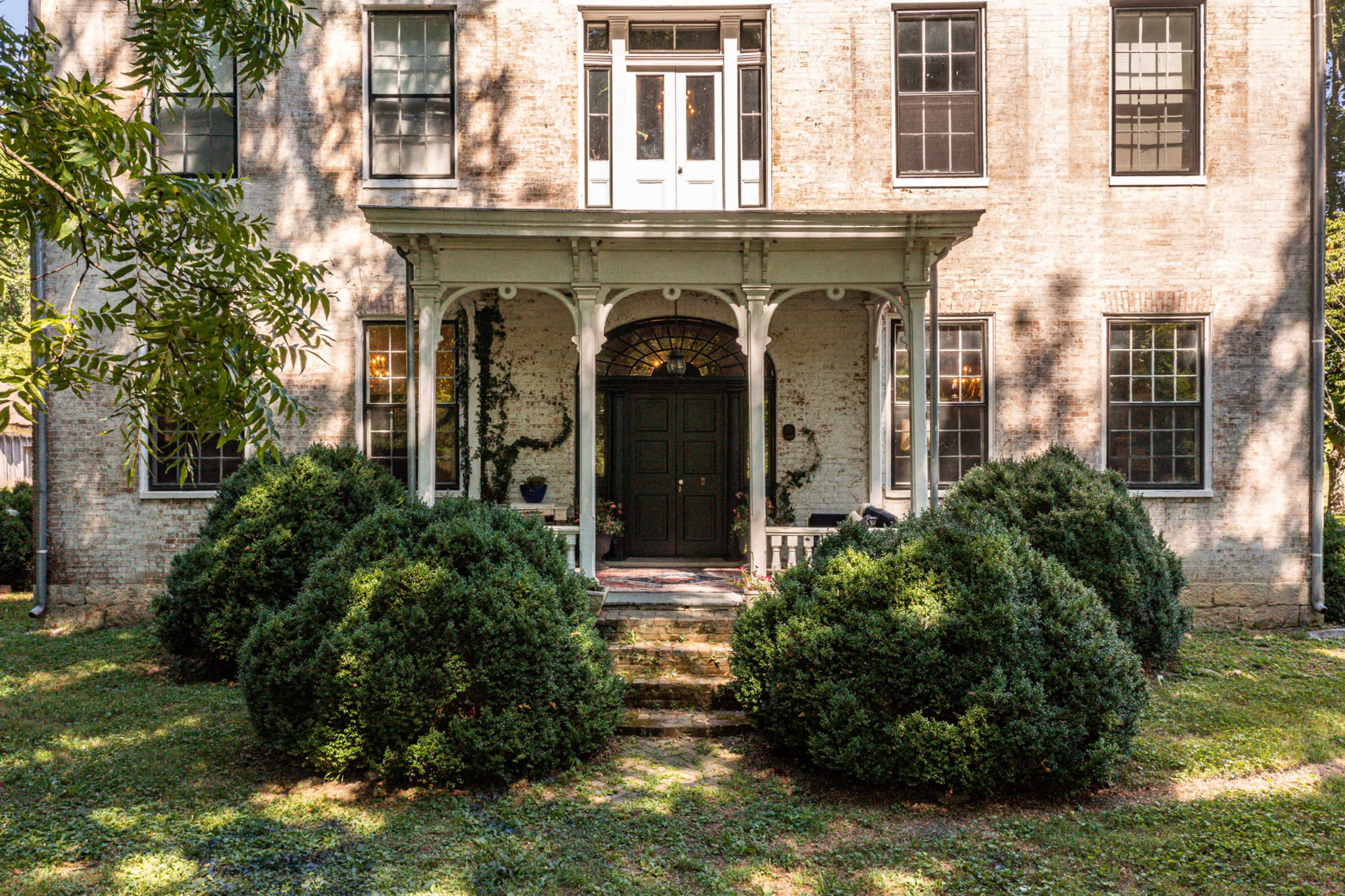 A historic two-story house with a covered porch, surrounded by neatly trimmed bushes and a grassy lawn.