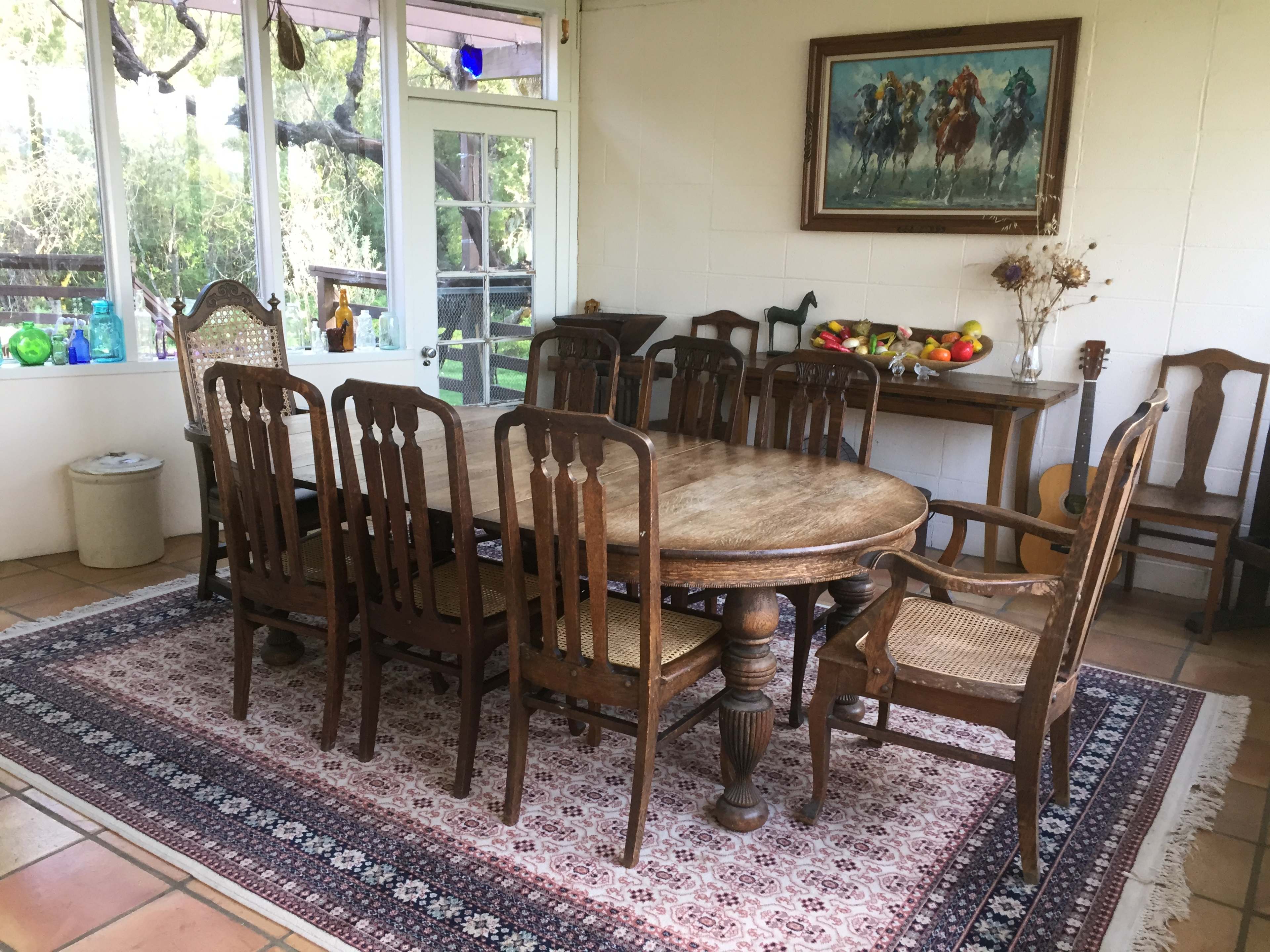 A large wooden dining table surrounded by chairs is set in a sunlit room with a rug, alongside a window that offers a view of greenery outside.