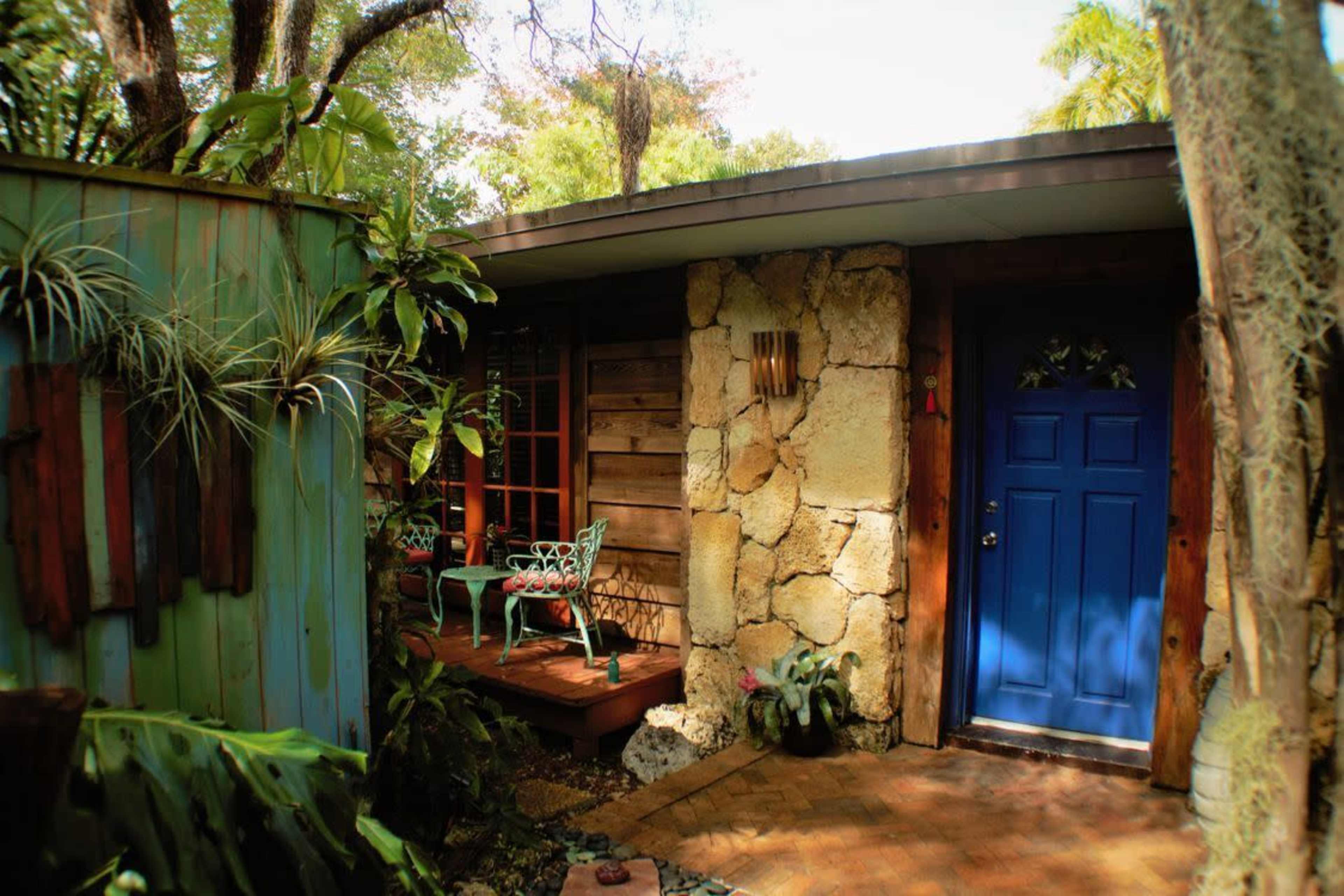 A small house with a blue door is surrounded by lush greenery and features a wooden deck with chairs.