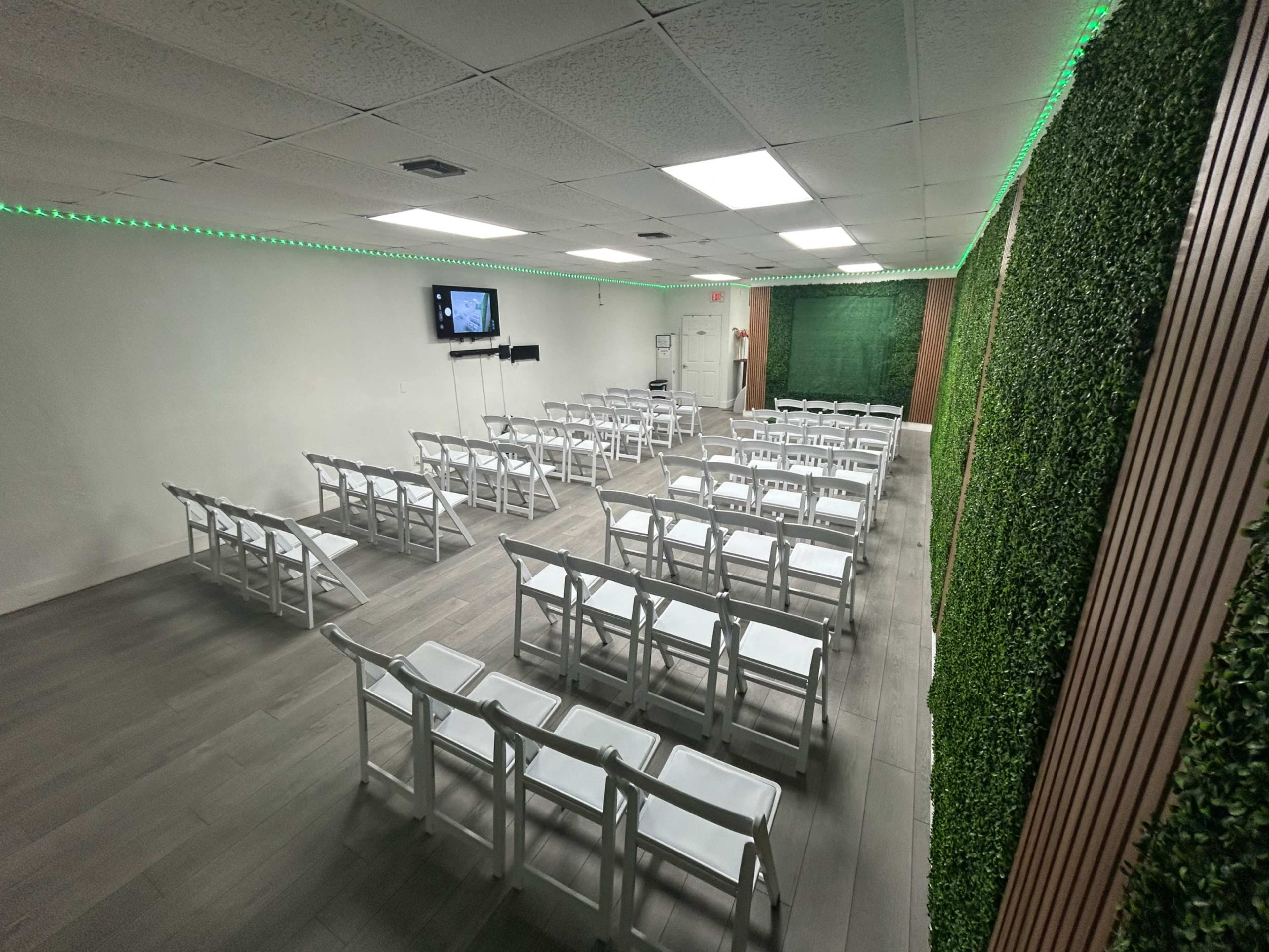 The image shows a room set up for an event, featuring rows of white folding chairs arranged in a well-lit space with green accent lighting and greenery on one wall.