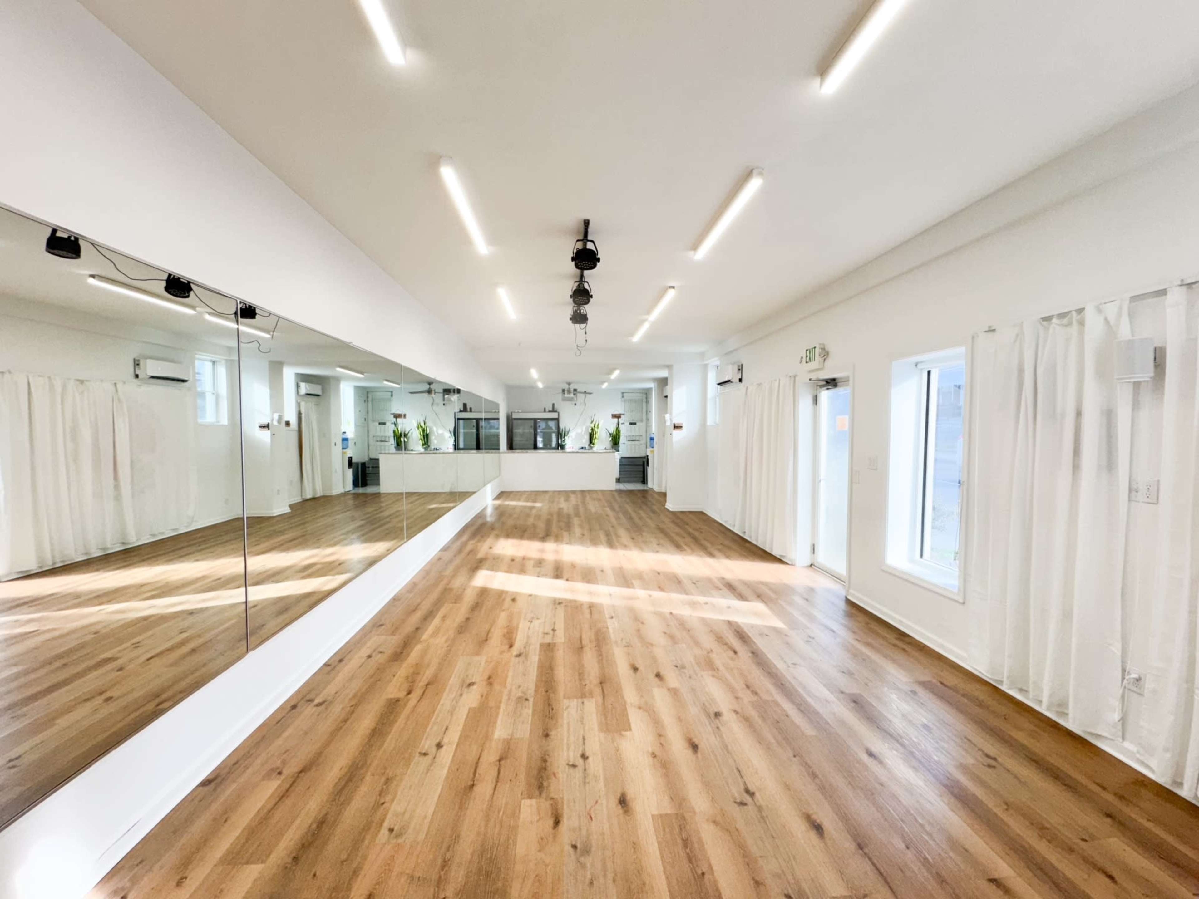 The image shows a spacious, well-lit studio with wooden flooring, a large mirror on one wall, and a kitchenette area in the background.