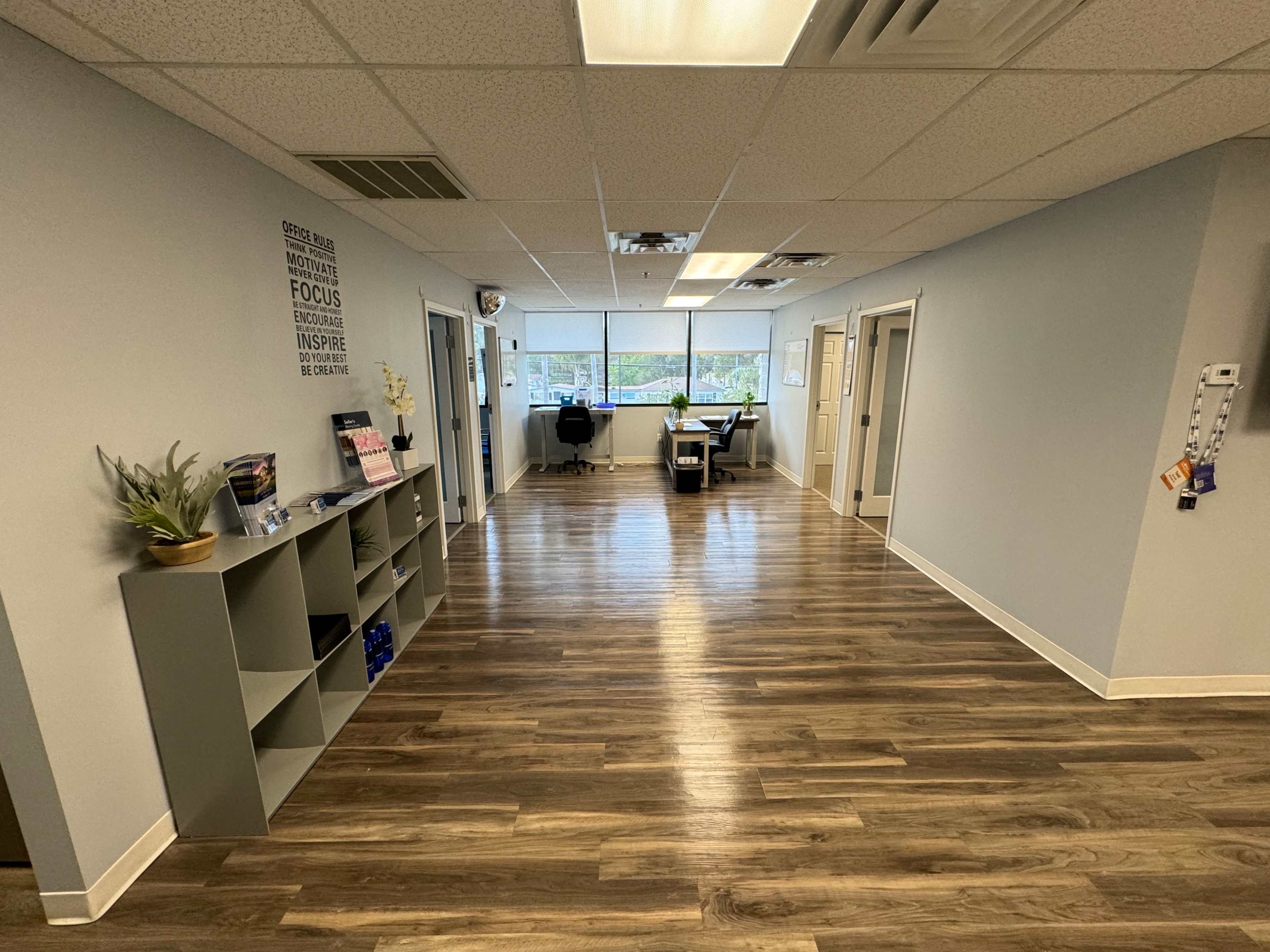 A well-lit hallway with grey walls, wooden flooring, and office doors leading to a workspace at the end.