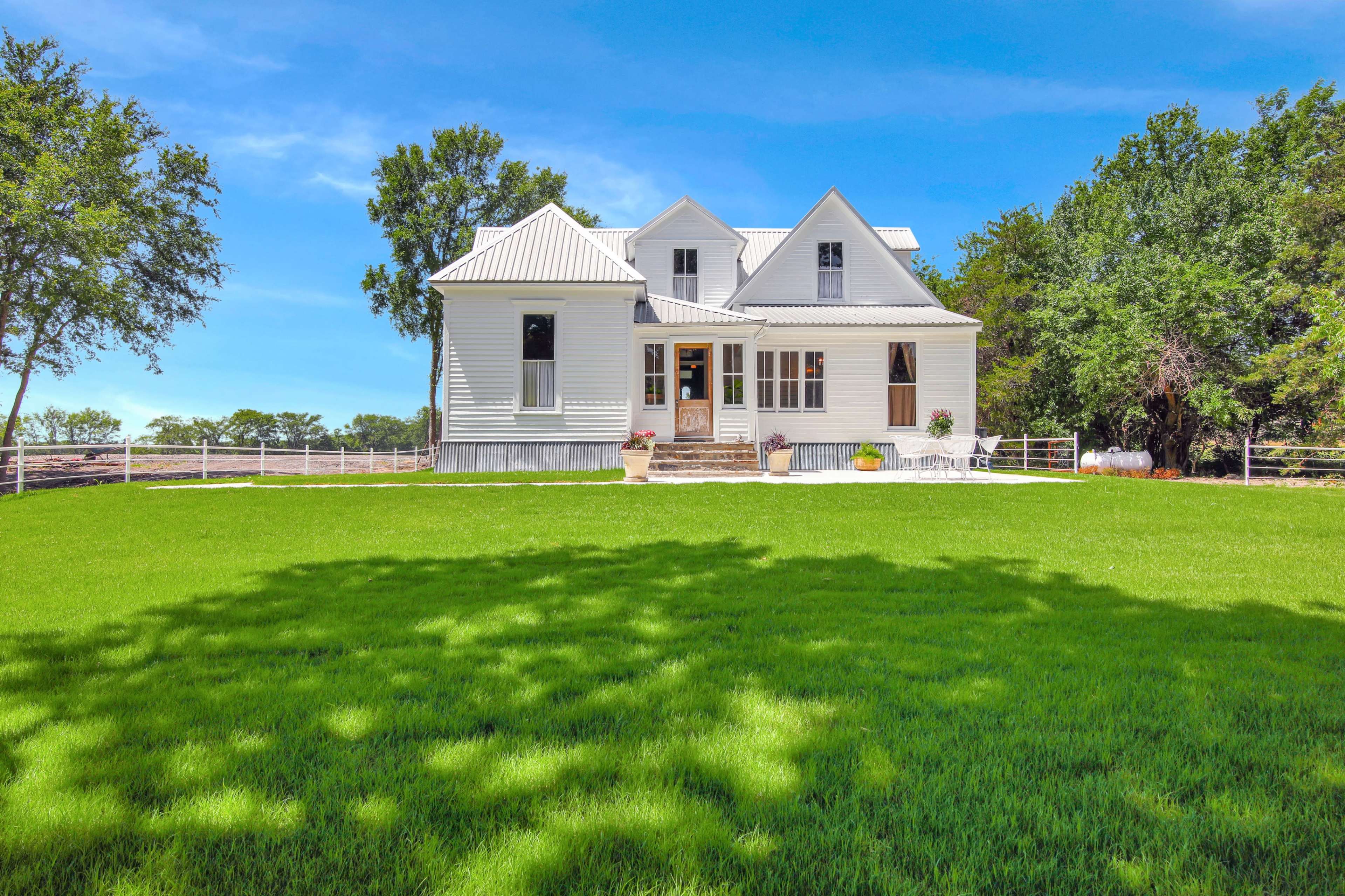 A white farmhouse with a metal roof sits on a well-kept lawn surrounded by trees.