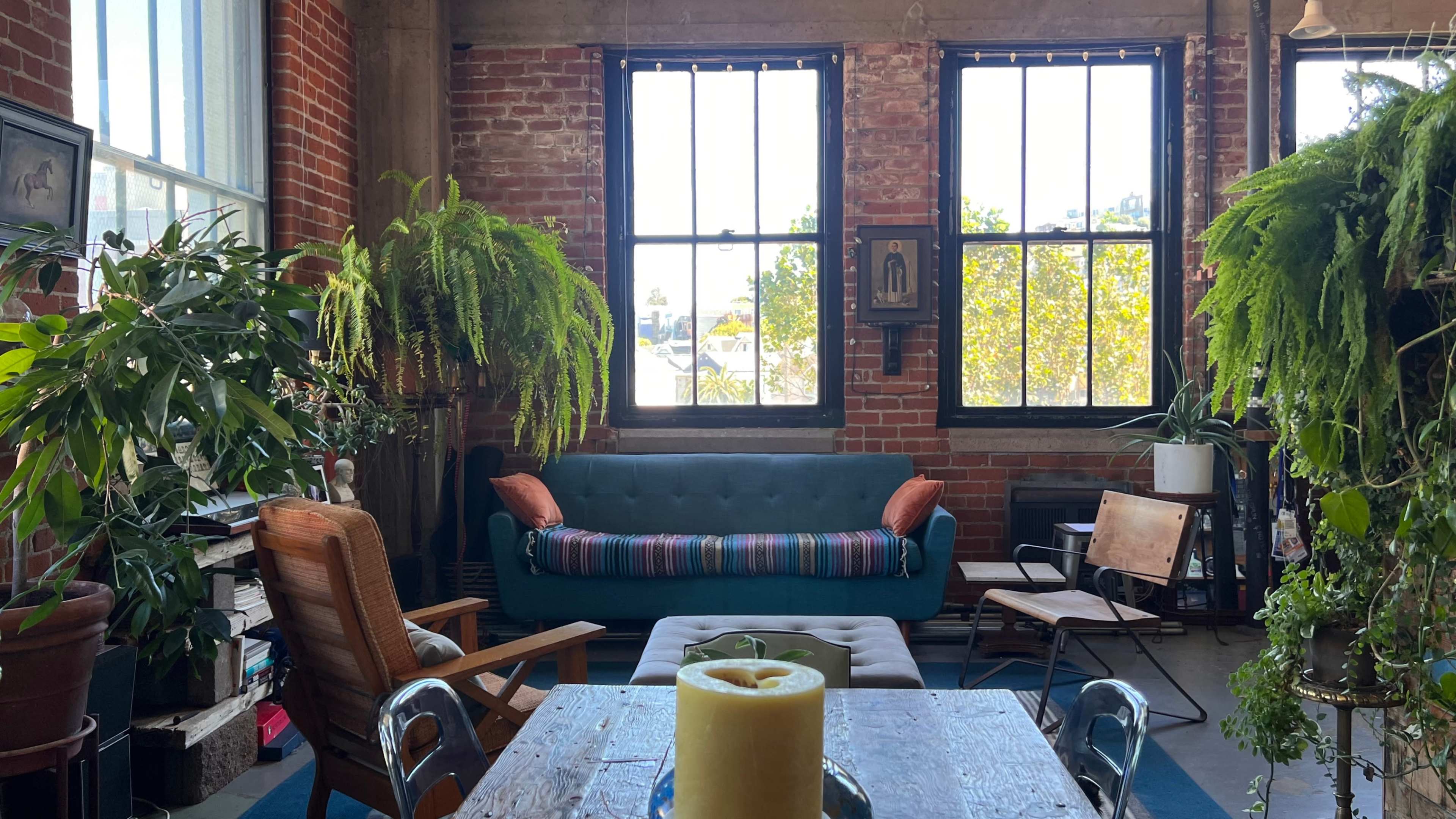 A cozy living room featuring a blue-striped sofa, wooden furniture, and various plants, with large windows letting in natural light.