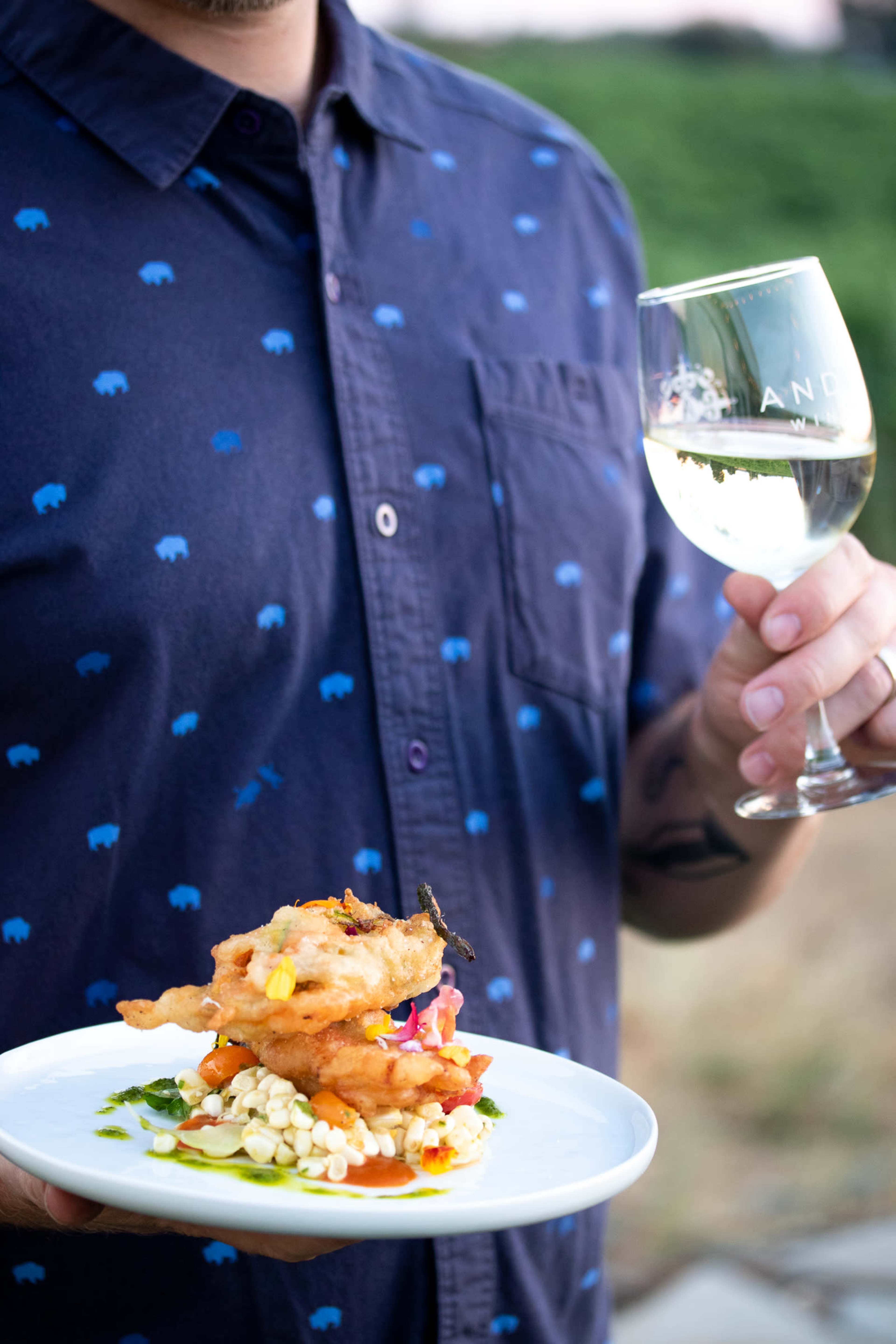 A person holds a plate of fried food atop a colorful vegetable base in one hand and a glass of white wine in the other.