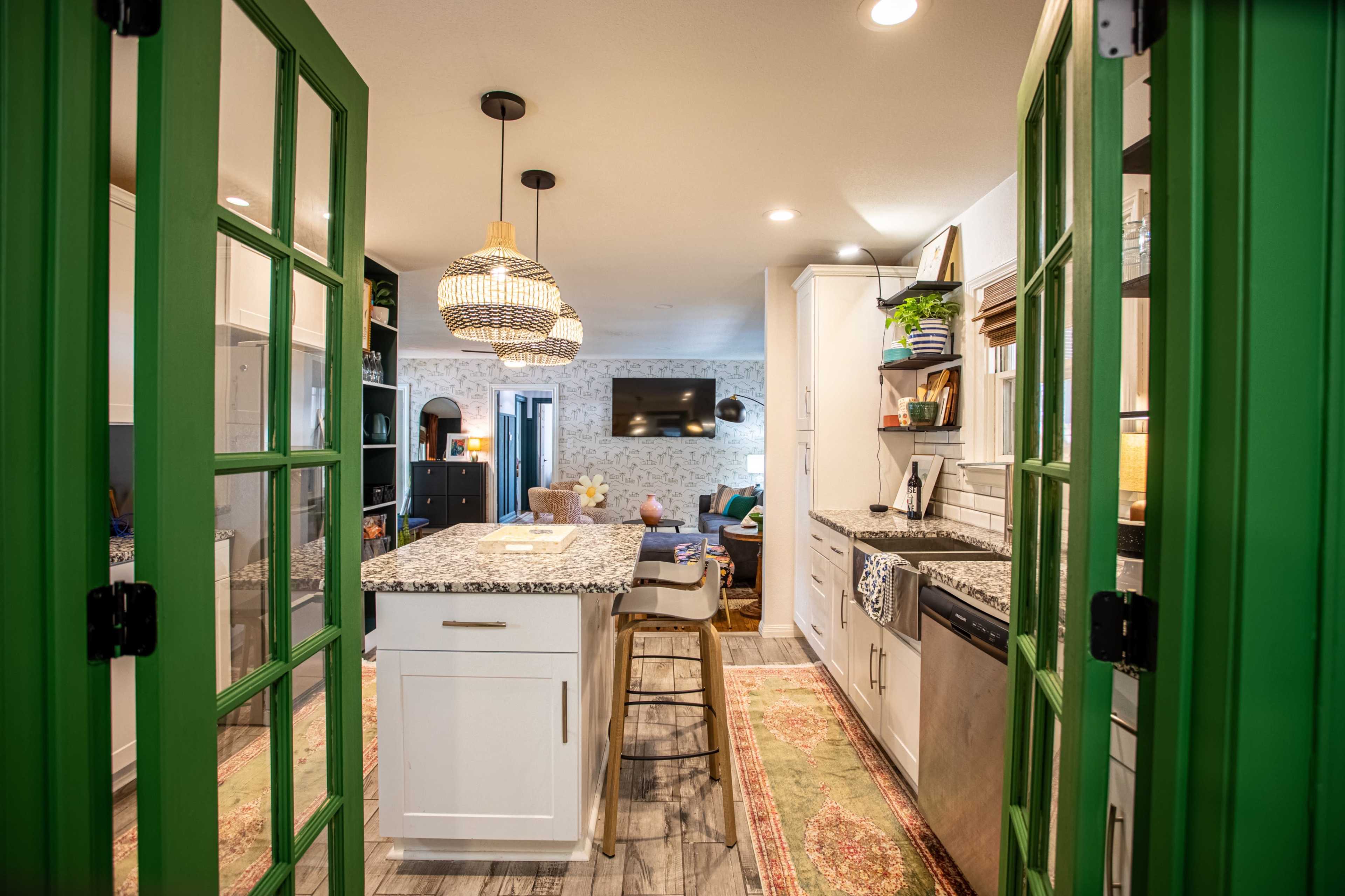 A modern kitchen viewed through green-framed double doors, featuring white cabinetry, a central island with bar seating, and a pendant light above.