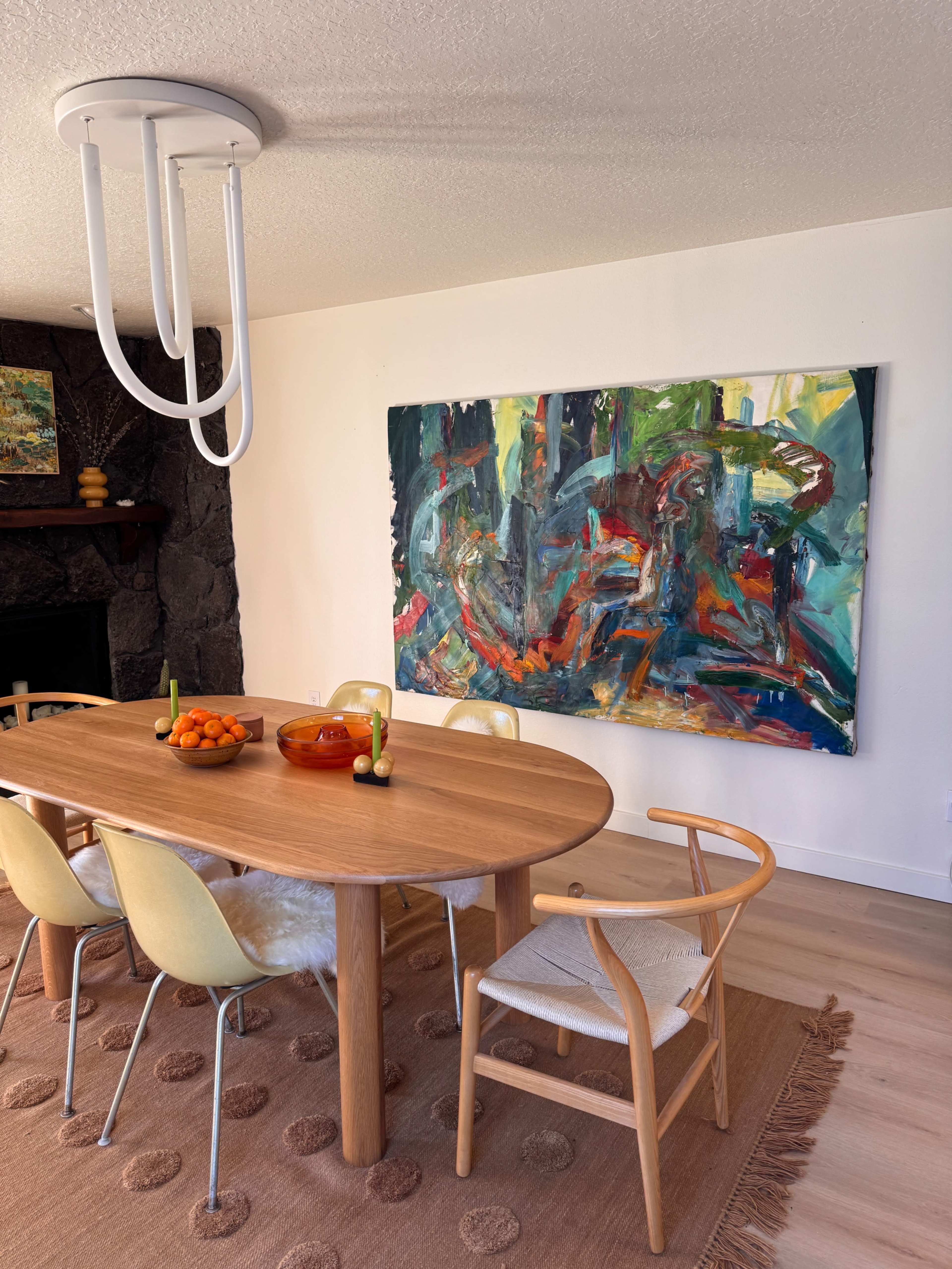 A wooden dining table with yellow chairs is set in a room featuring a large abstract painting on the wall and a round light fixture above.