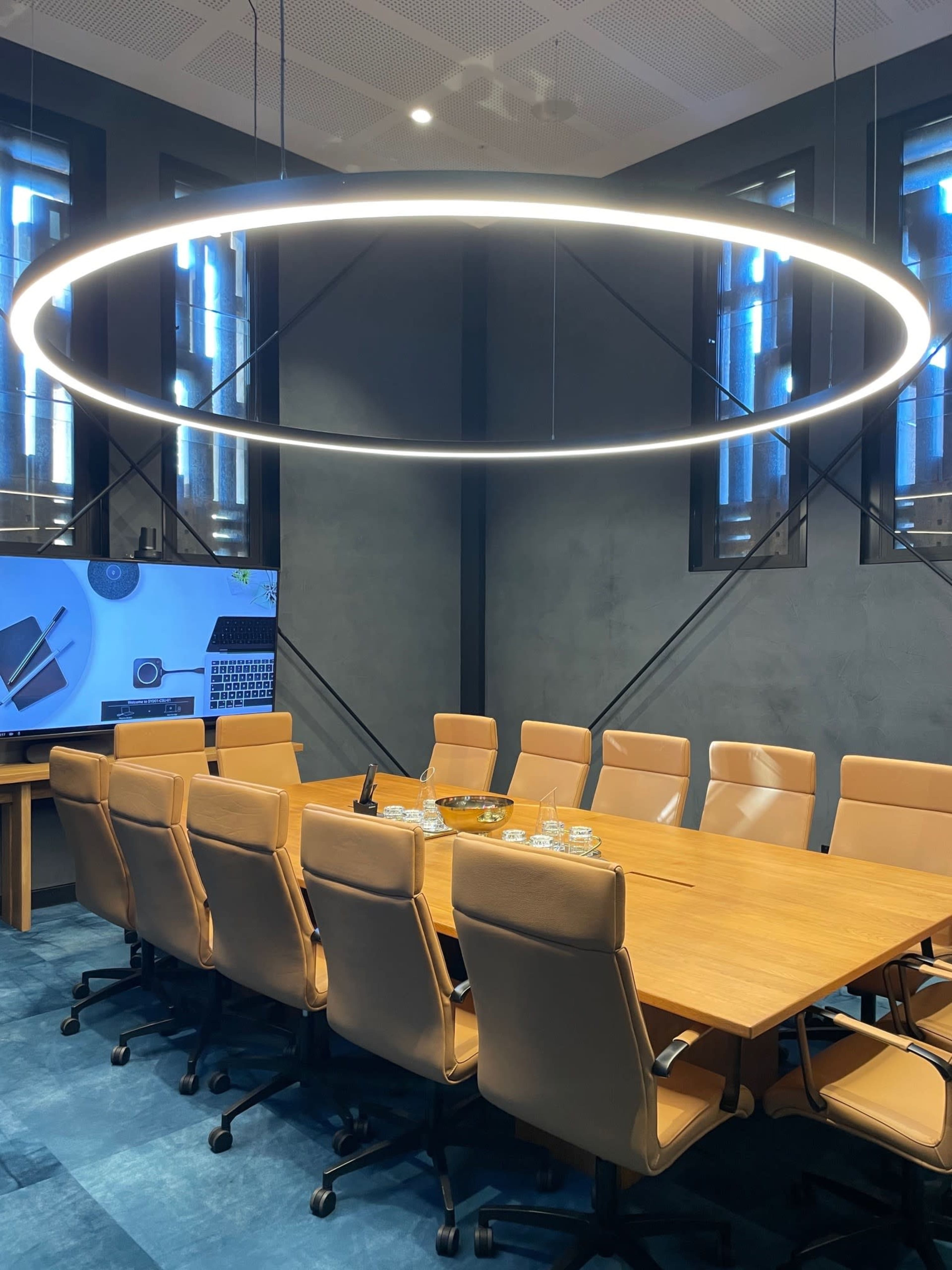 A large conference room features a rectangular wooden table surrounded by beige chairs, with a circular light fixture hanging above and a screen displaying technology-related content.