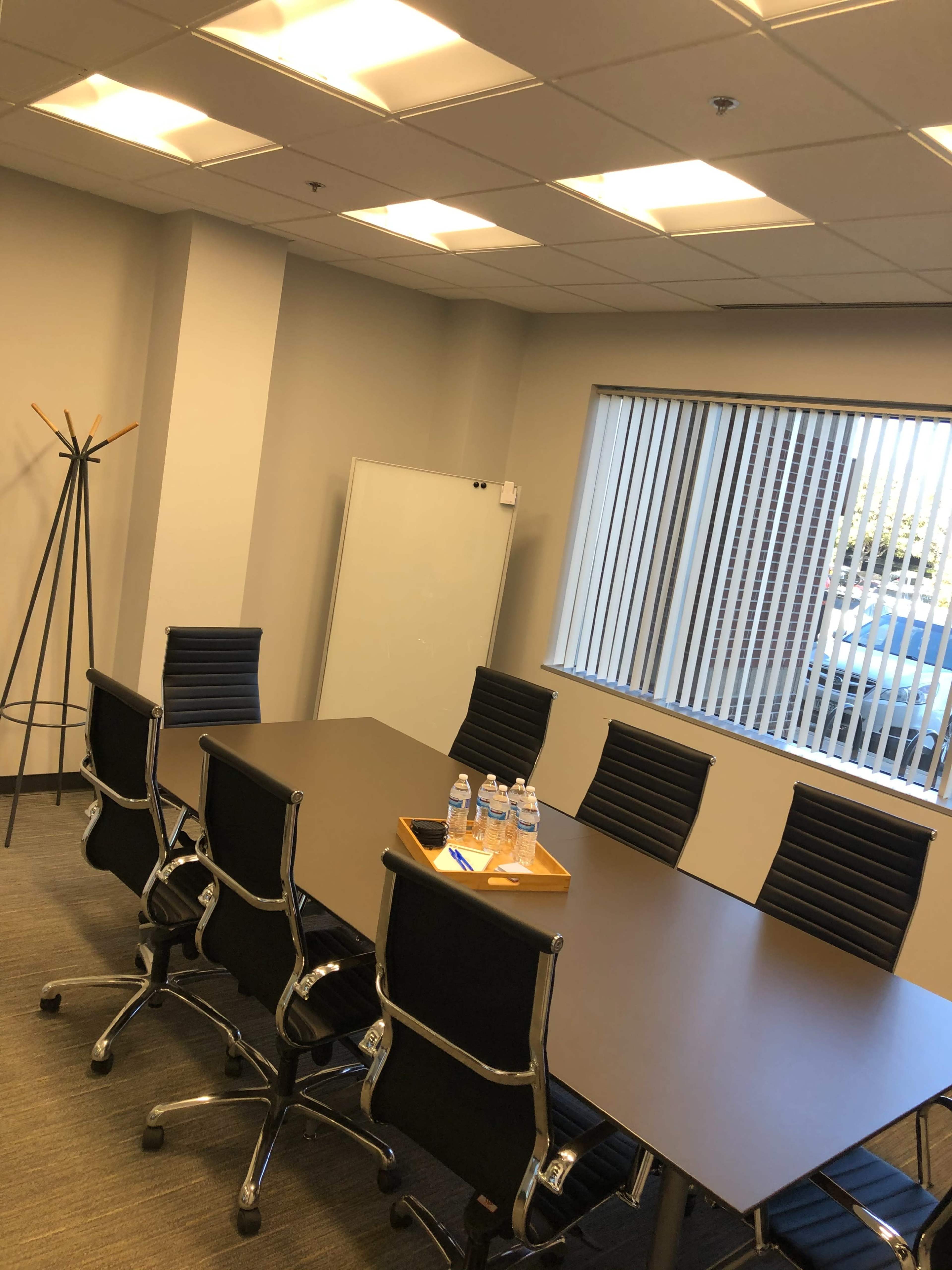 A conference room features a large table surrounded by black chairs, a coat stand in the corner, and several water bottles on a tray.