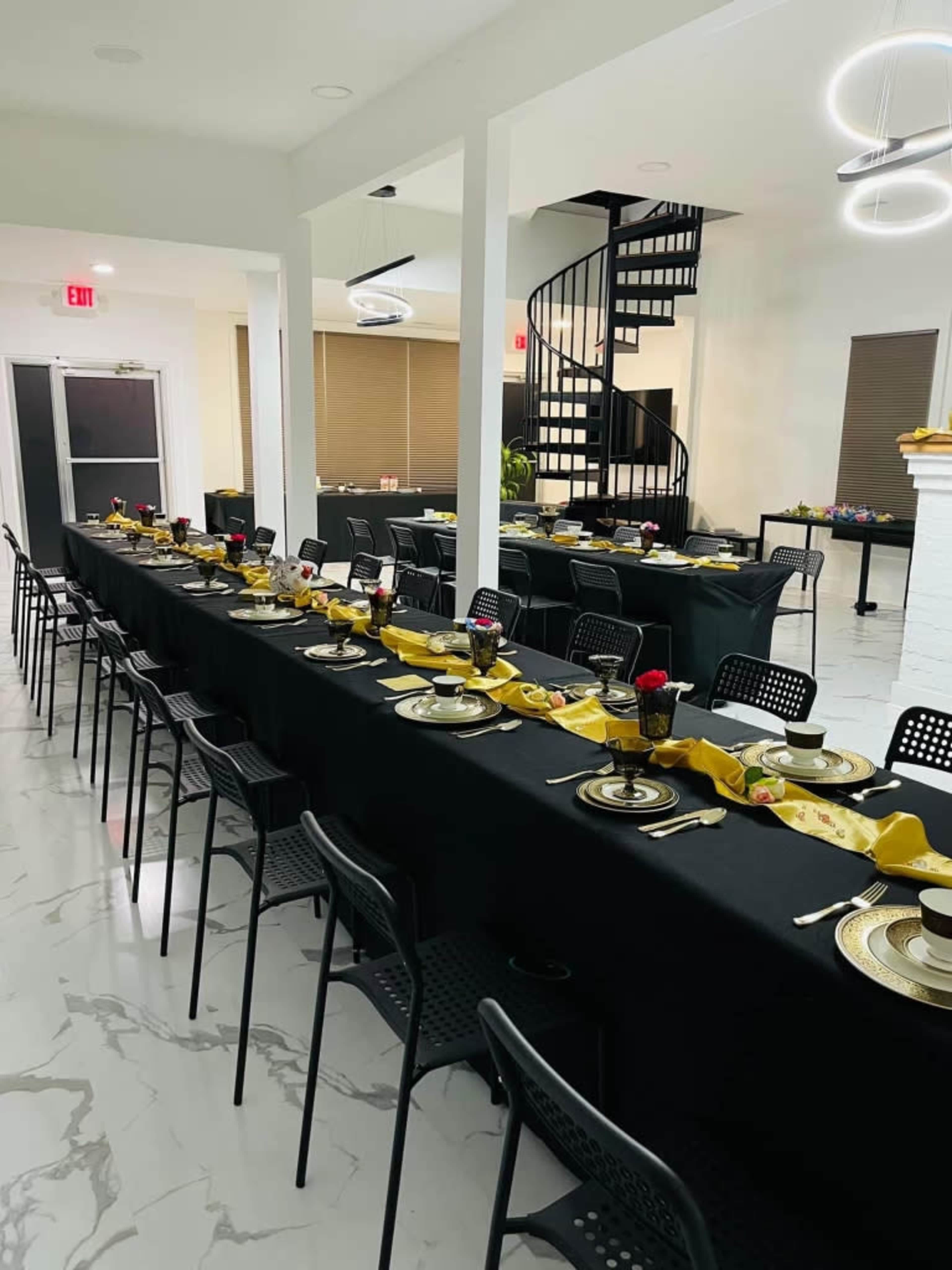 The image shows a large dining room with several long tables set with black tablecloths, plates, and floral centerpieces, and a spiral staircase visible in the background.