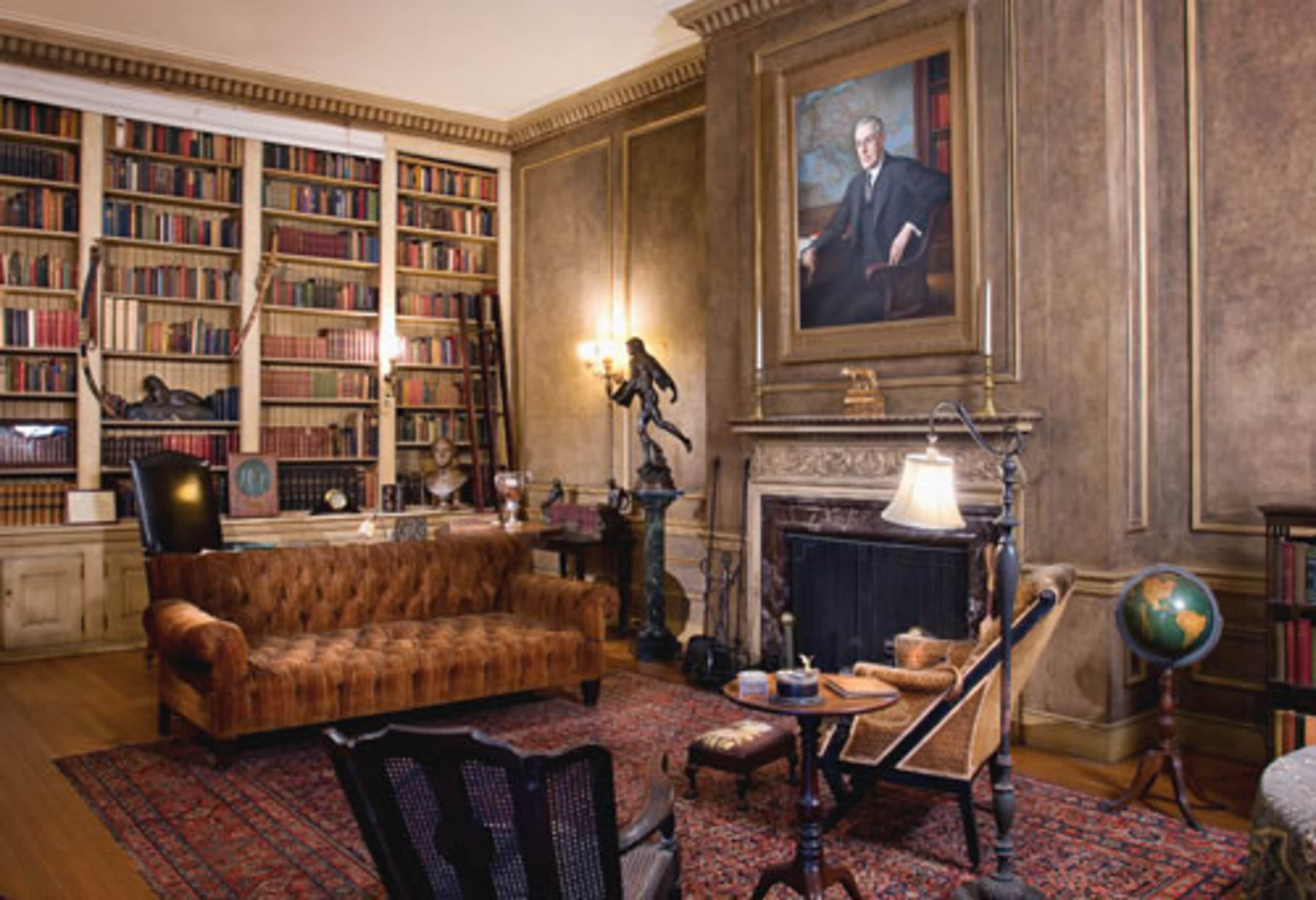 A well-furnished library with a leather sofa, bookshelves filled with books, a portrait on the wall, and a fireplace.