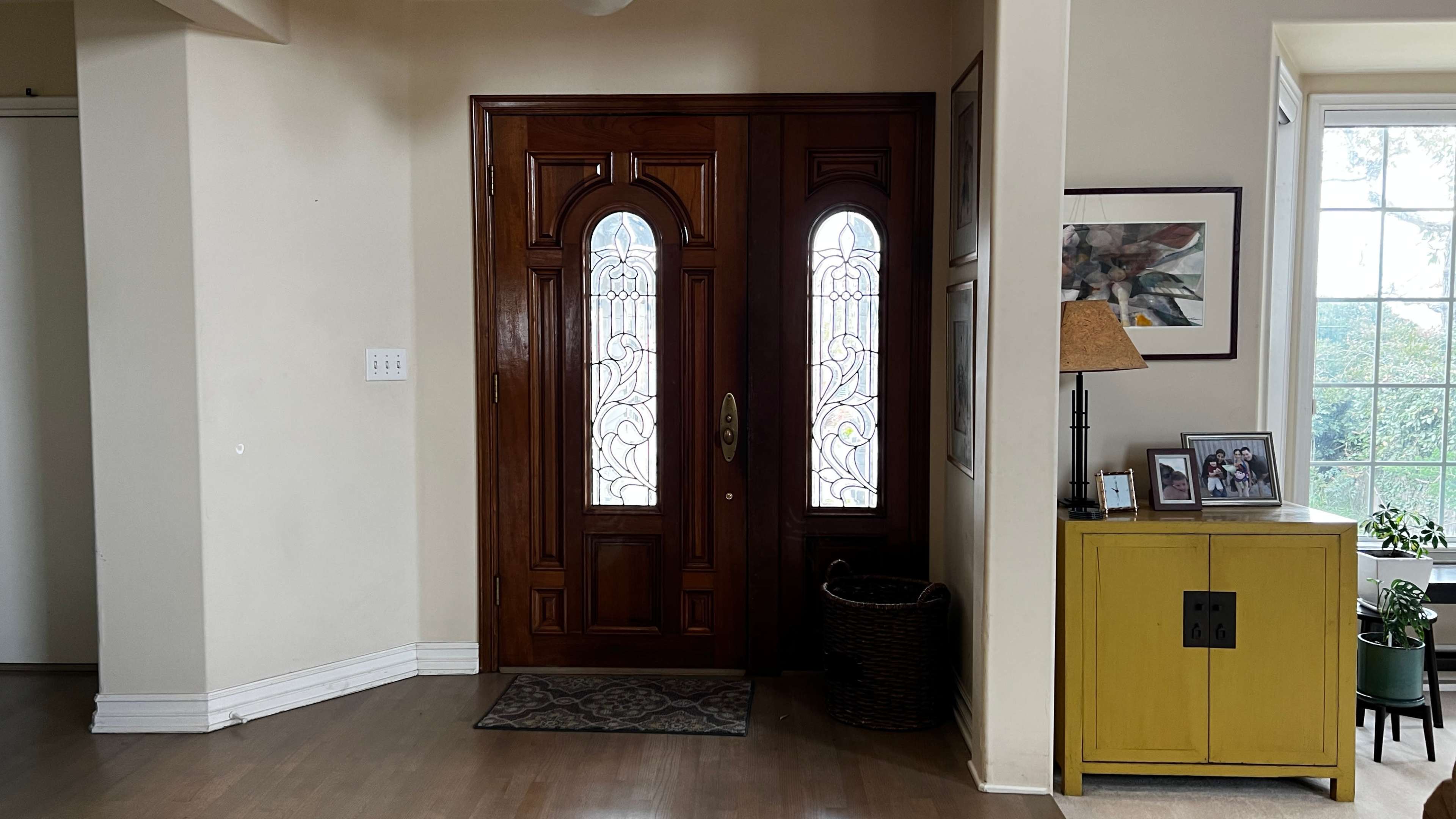 A spacious entryway with a pair of wooden double doors featuring stained glass panels, a decorative rug, and a yellow cabinet next to a lamp and framed photographs.