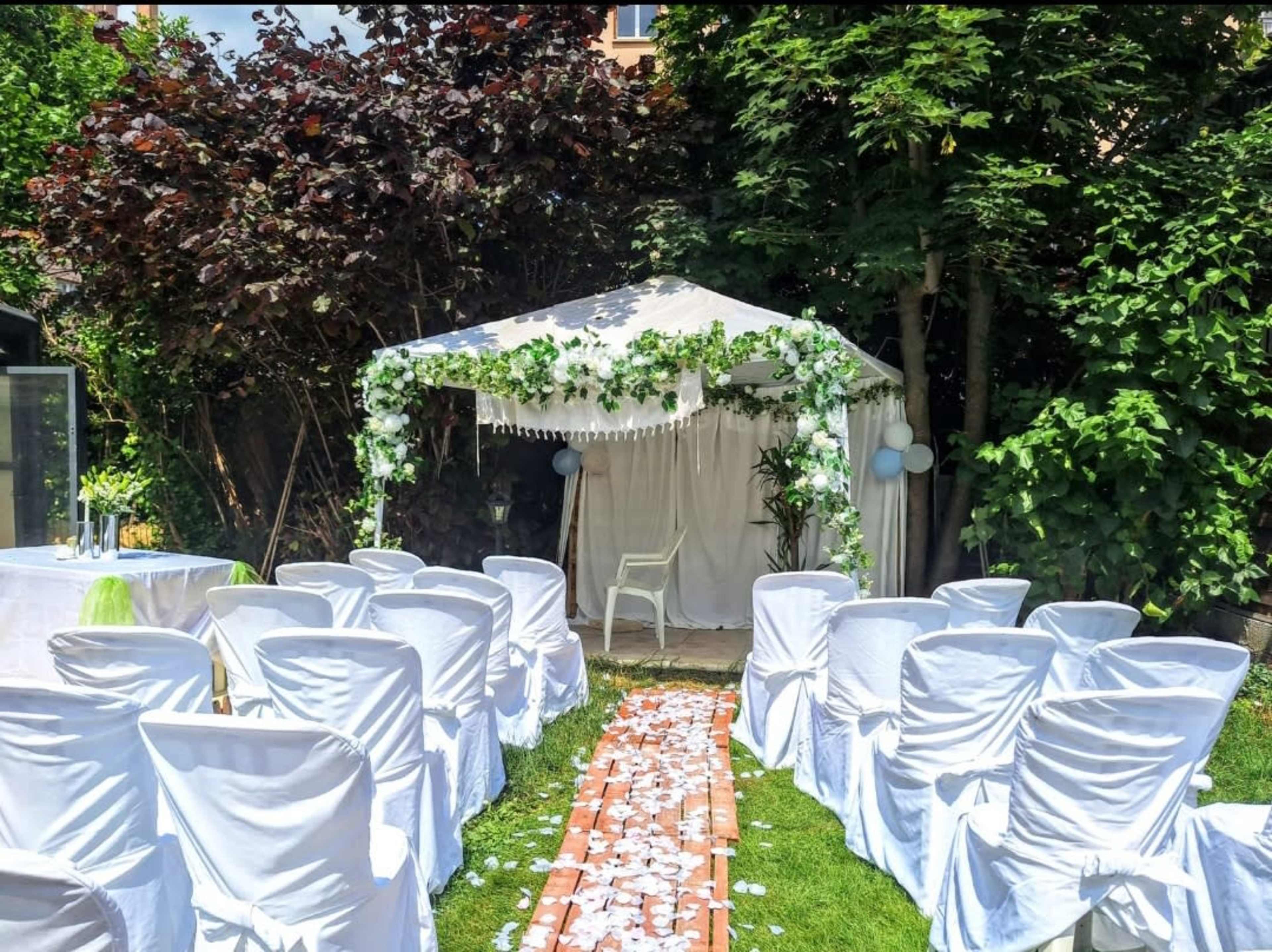 The image shows a garden wedding setup with white chairs arranged in rows facing a decorated gazebo surrounded by greenery.