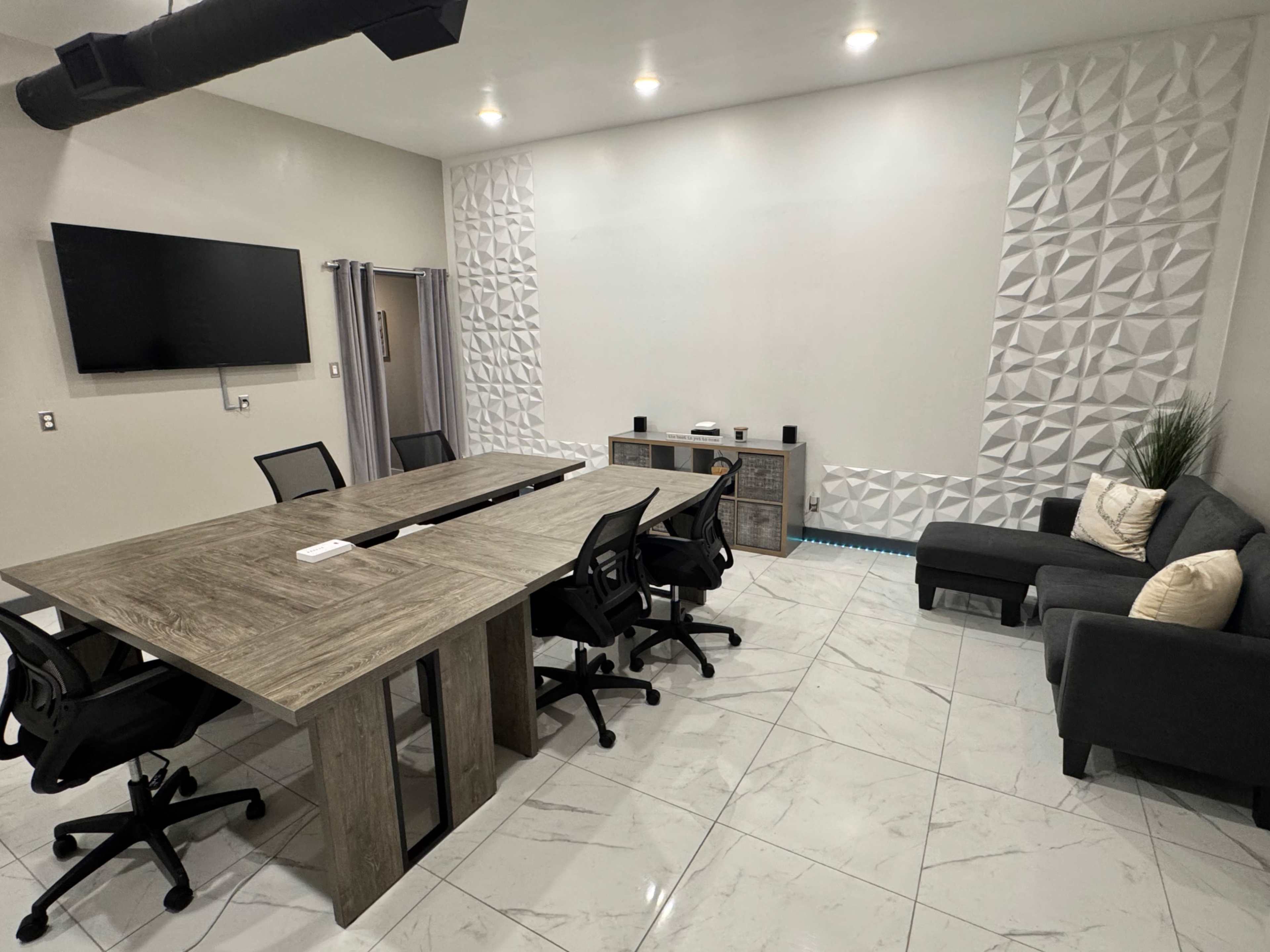 The image shows a modern conference room with a large wooden table surrounded by black office chairs, a large wall-mounted screen, and a seating area with a gray couch and decorative elements.