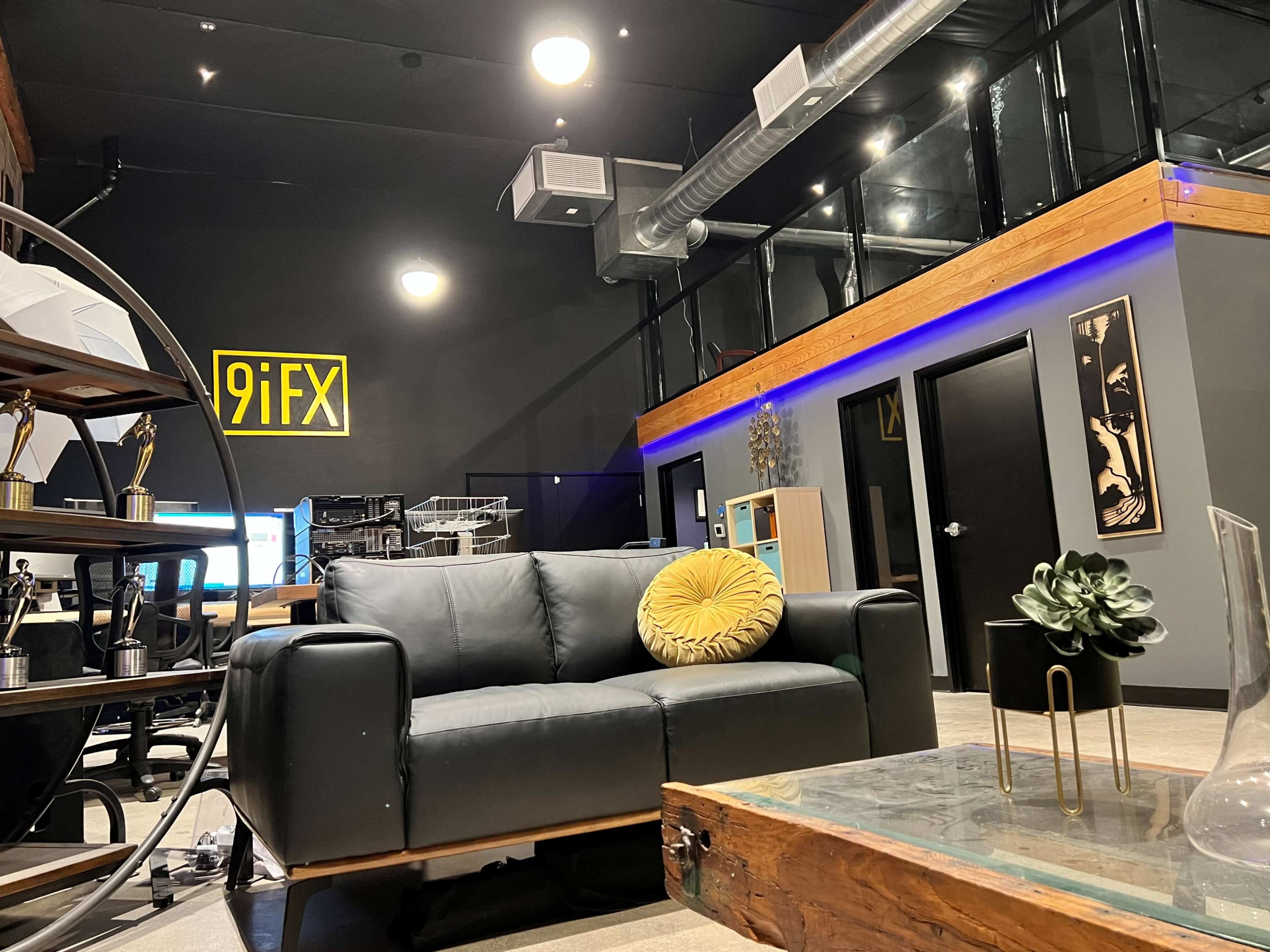 The image shows a modern office space featuring a black couch with a yellow pillow, a wooden coffee table, and a staircase leading to a mezzanine level with blue lighting.