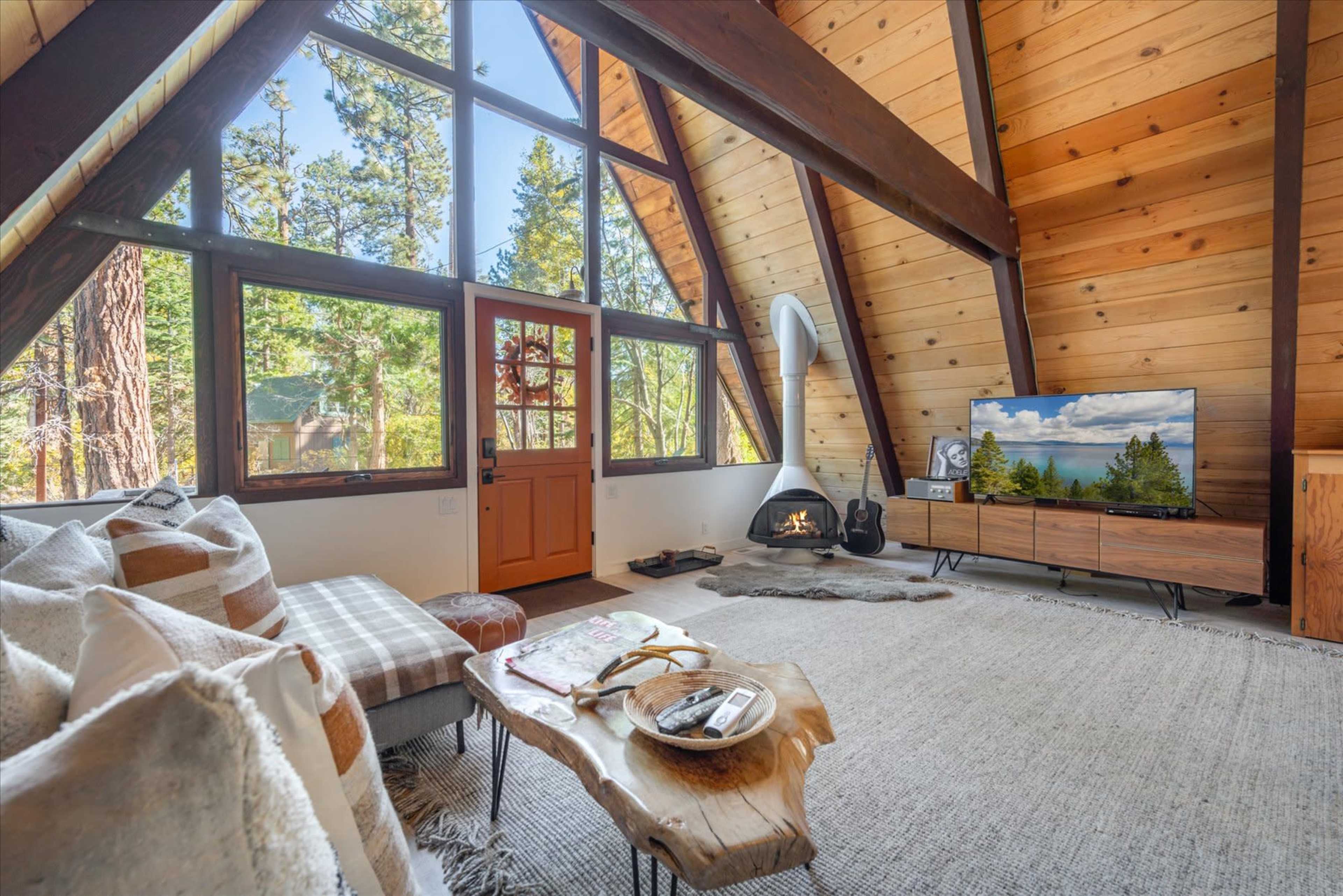 A cozy living room features a large window with natural light, wooden beams, and a modern TV setup beside a small fireplace.