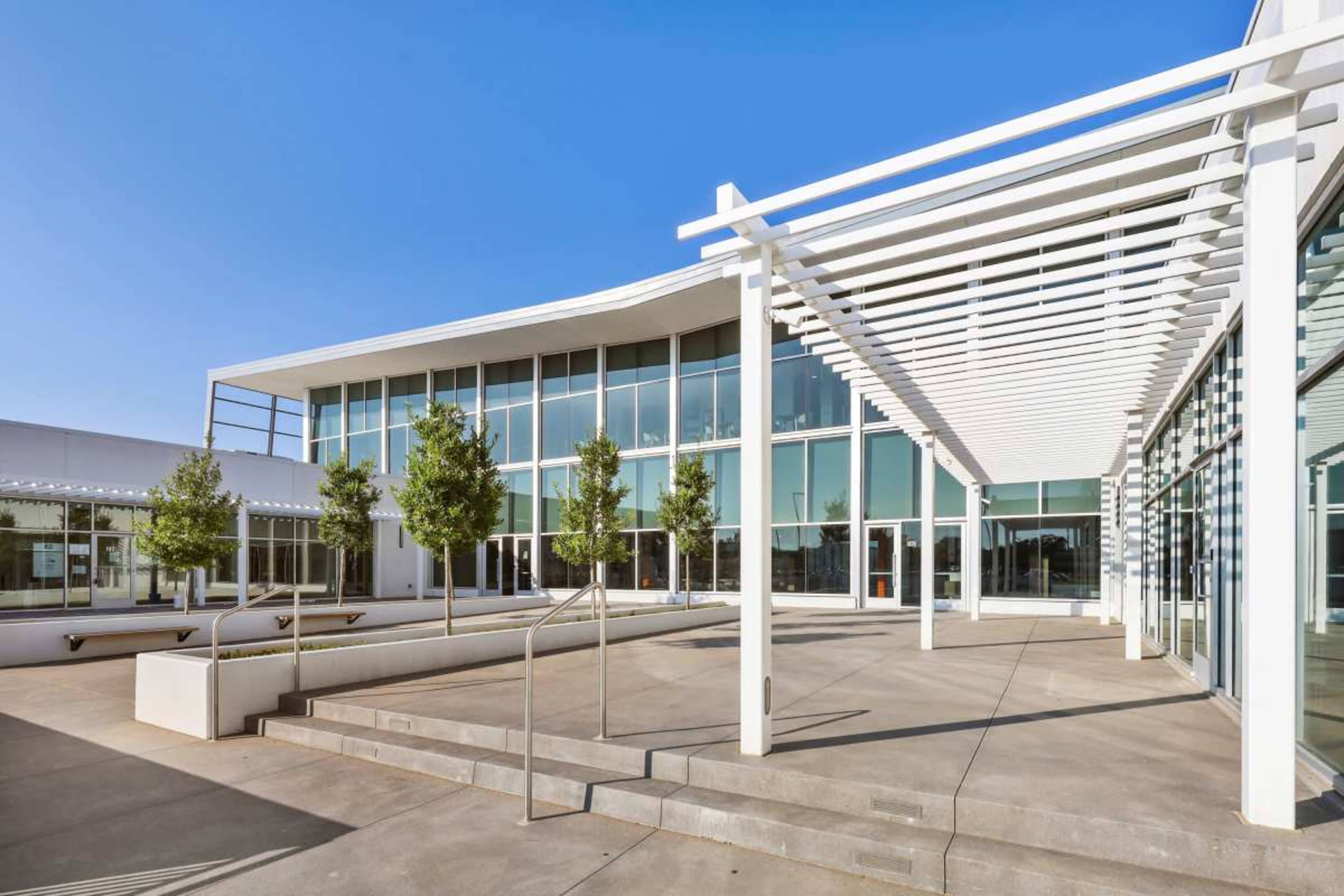 The image shows a modern building with large glass windows, surrounded by trees and a paved outdoor area under a white pergola.