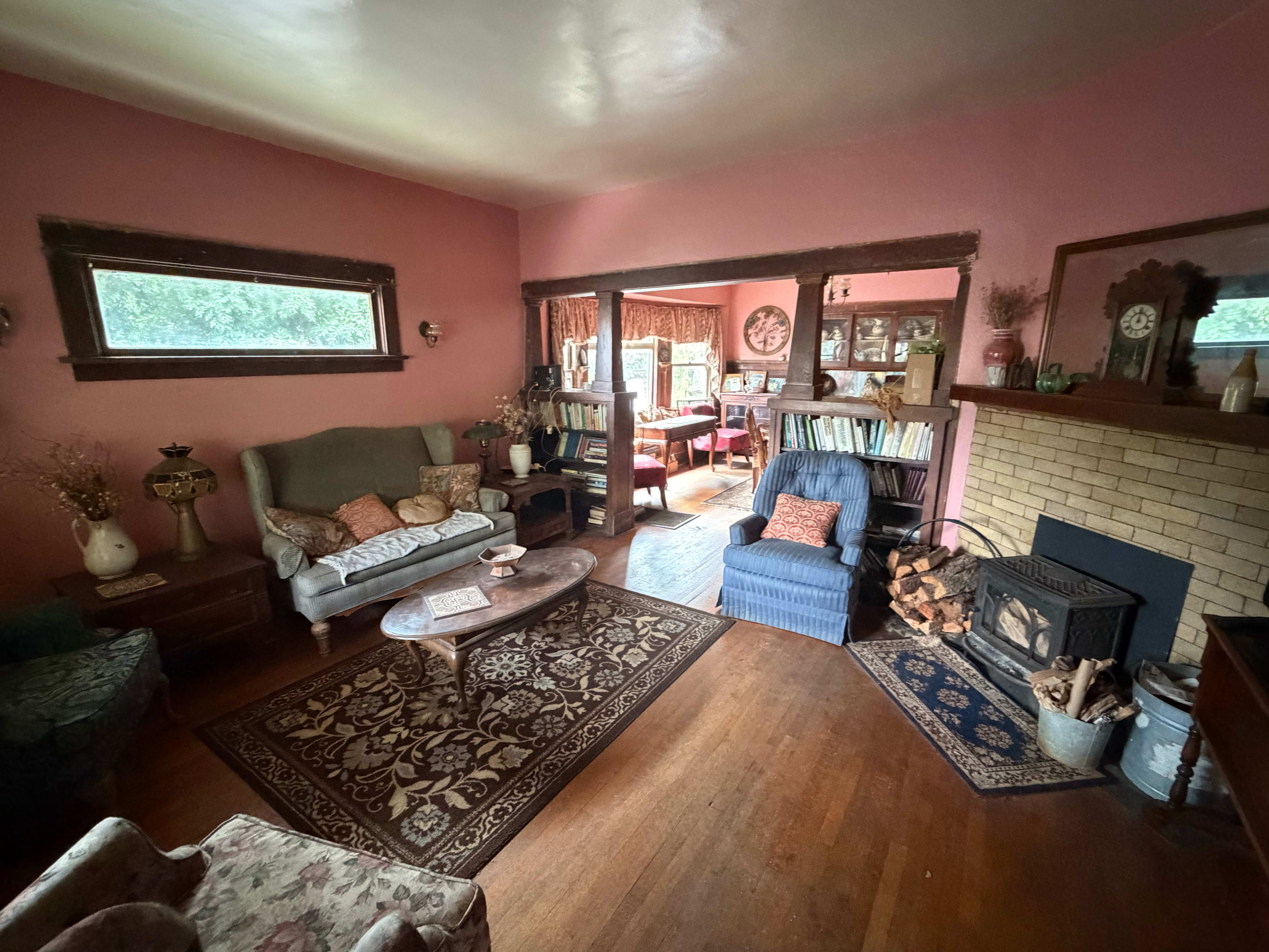 The image shows a cozy living room with pink walls, a fireplace, multiple seating options, and a view into a dining area filled with bookshelves.