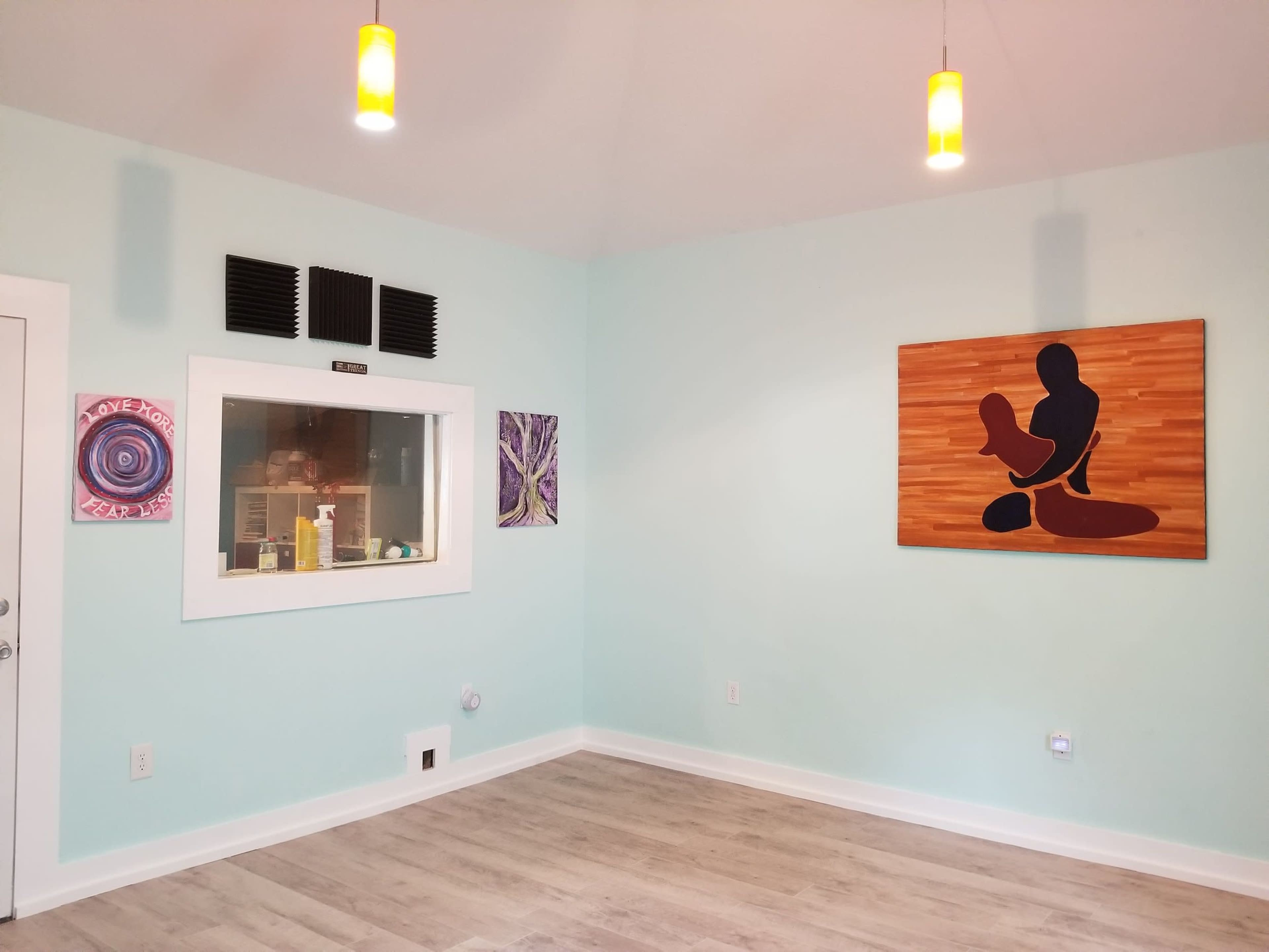 A light blue room with wooden flooring, featuring a large painting on the right wall, two framed artworks, and a glass-walled booth on the left.