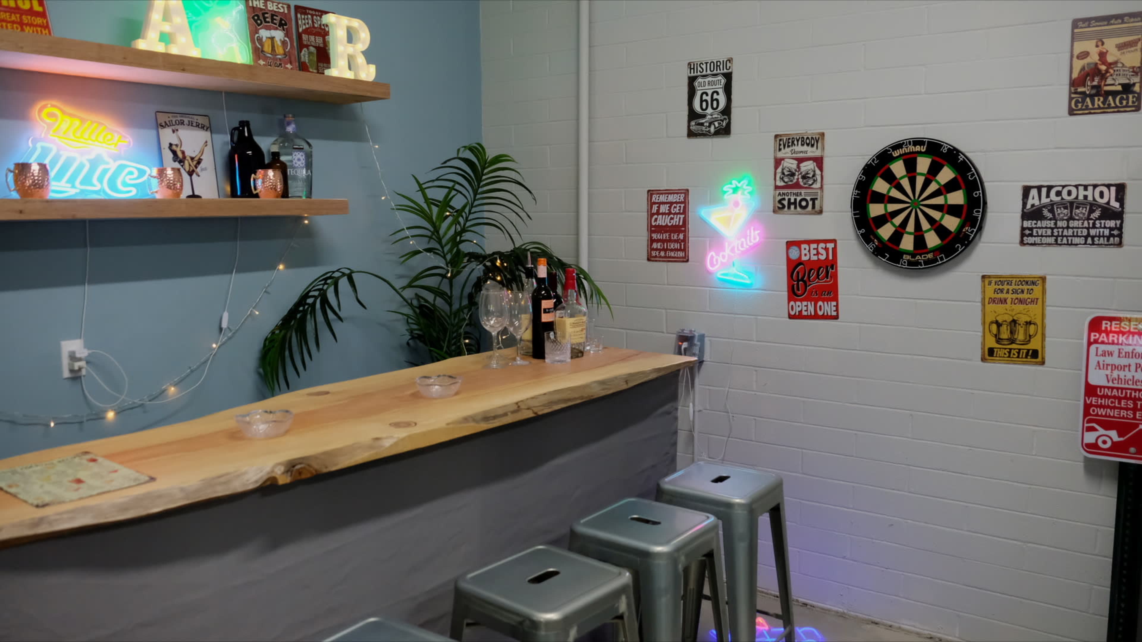 A small home bar features a wooden counter with metal stools, shelves displaying bottles, and various vintage signs, alongside a dartboard and plants.