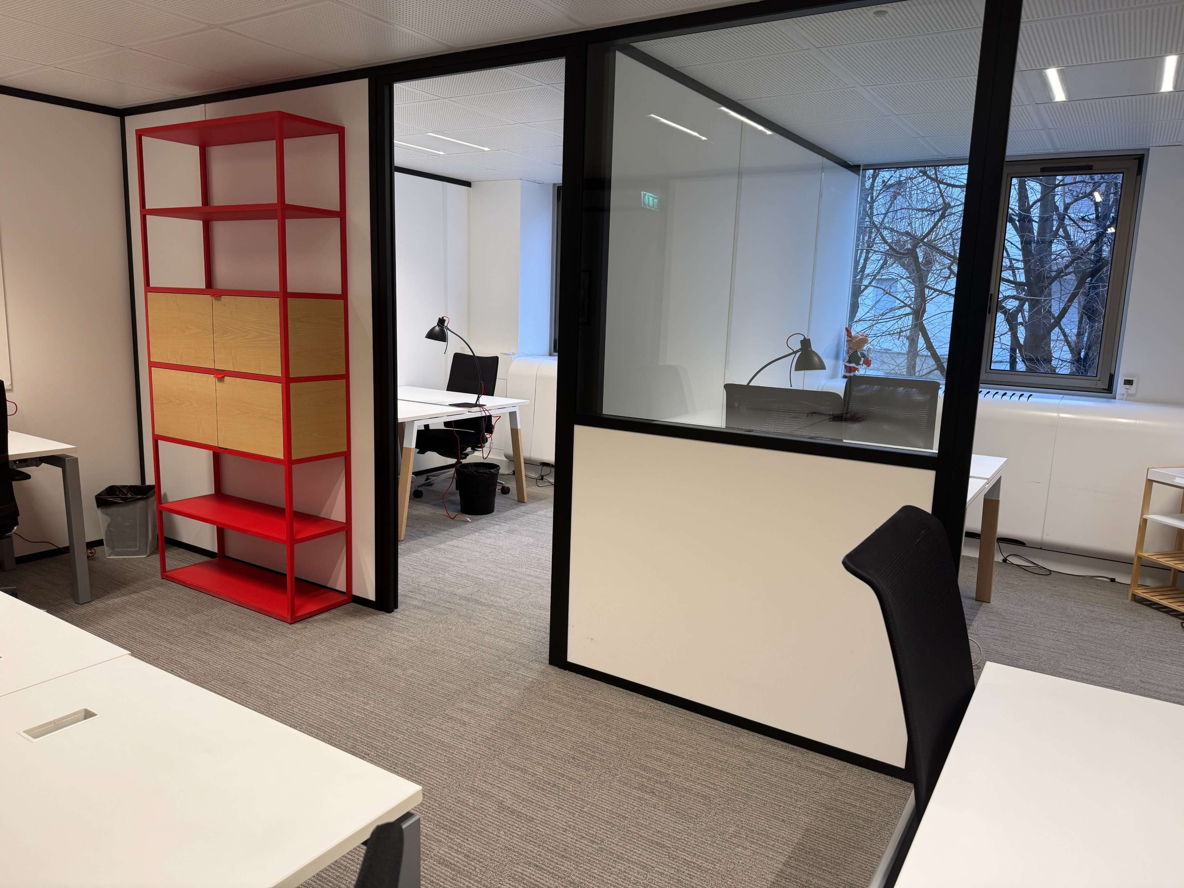 The image shows an office space divided by glass walls, featuring desks, a bookshelf, and large windows with trees visible outside.