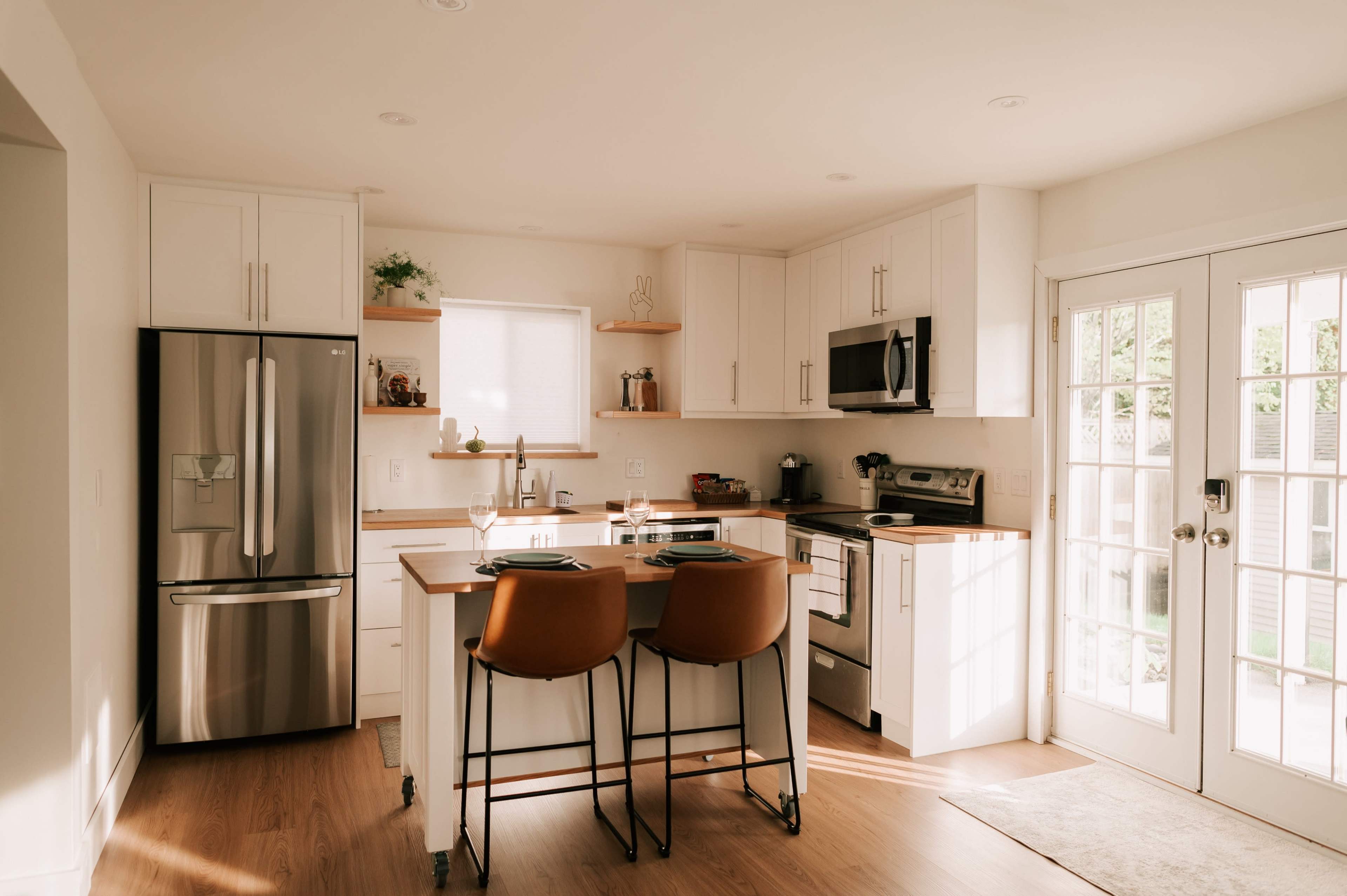 The image shows a modern kitchen featuring white cabinets, stainless steel appliances, a wooden island with two orange bar stools, and a door leading to an outdoor area.
