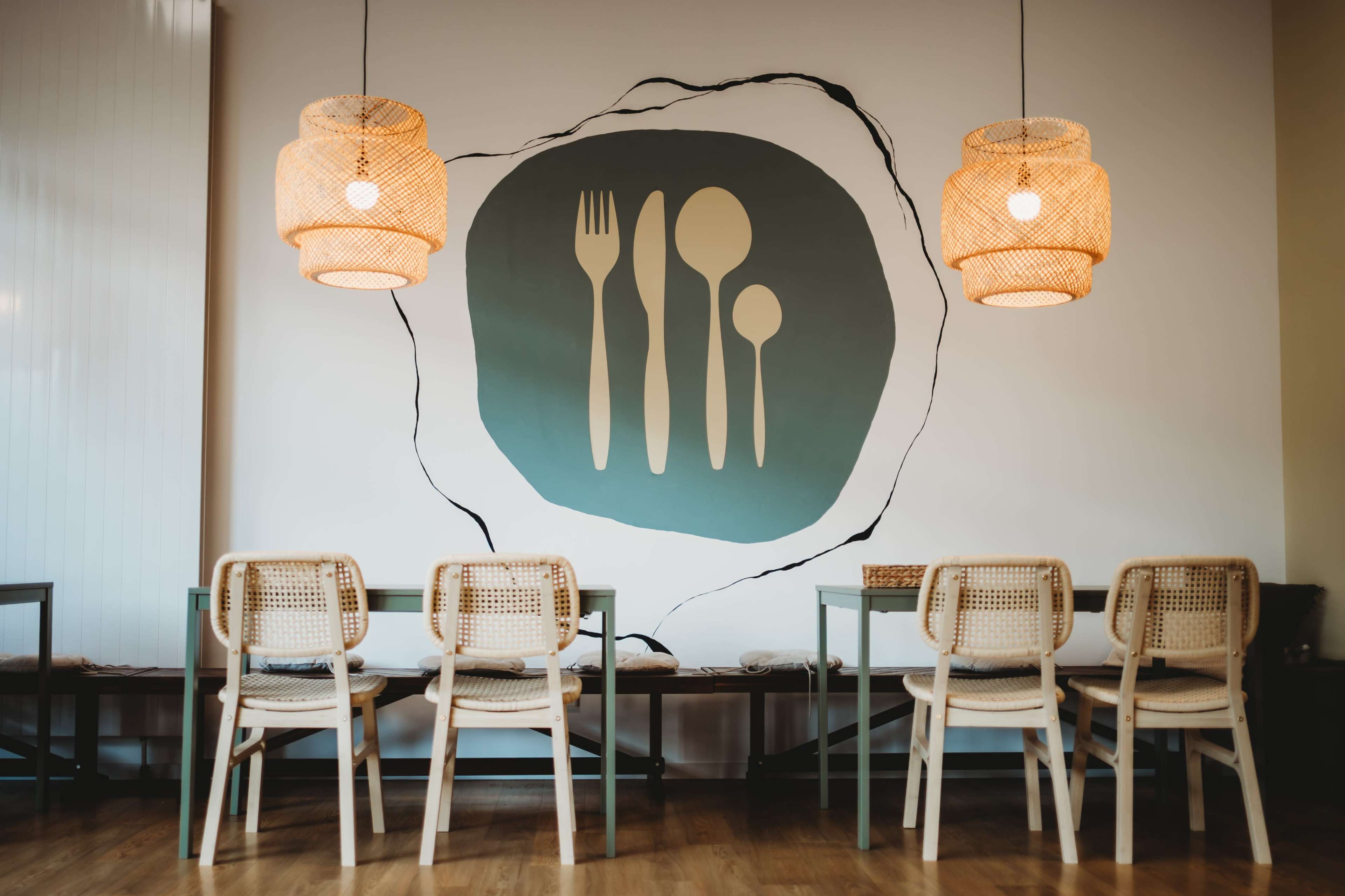 The image features a dining area with wooden chairs and a wall decor depicting cutlery against a light-colored backdrop, complemented by woven pendant lights.