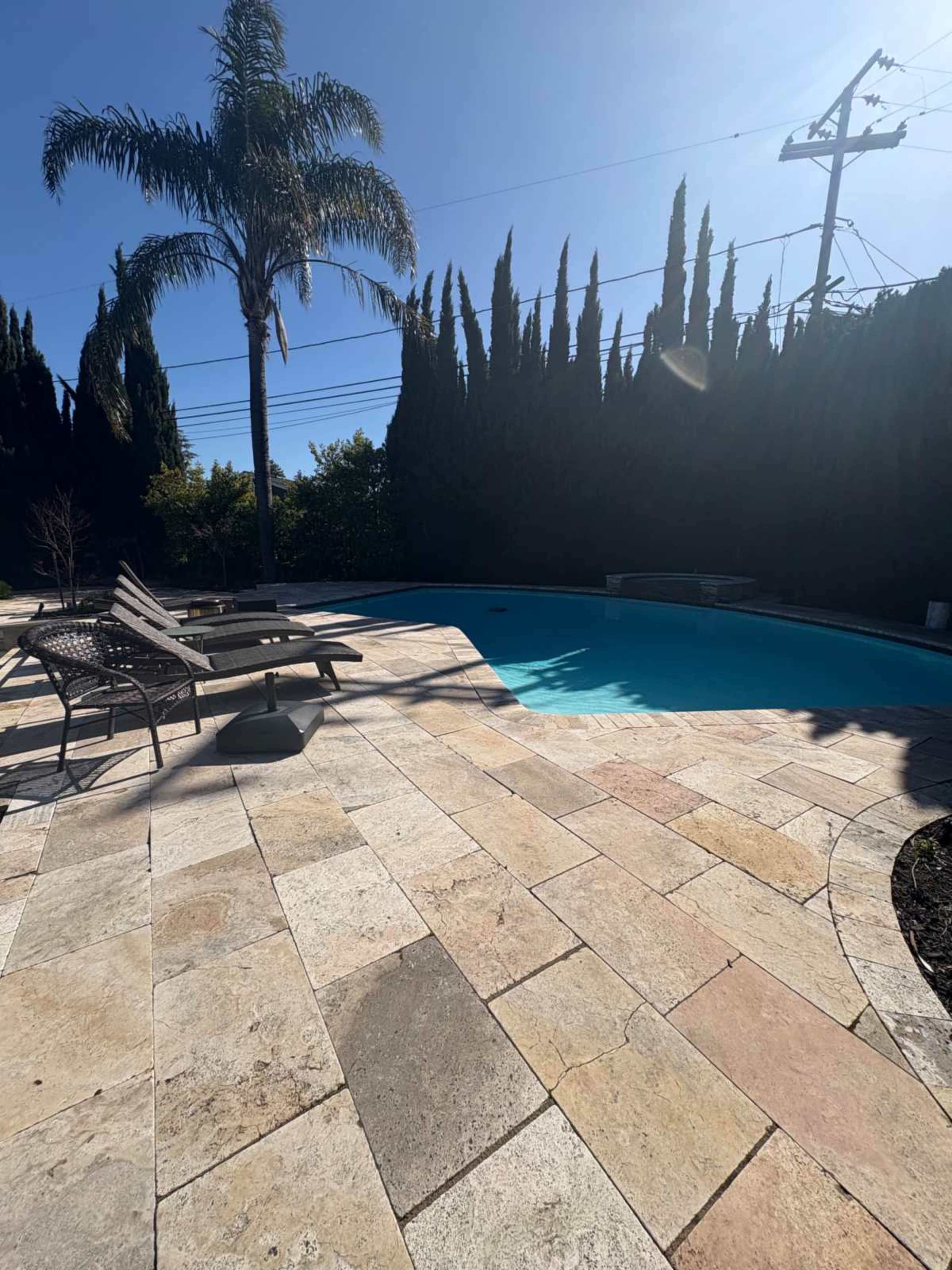 A clear blue swimming pool is surrounded by stone tiles and palm trees in a sunlit outdoor space.