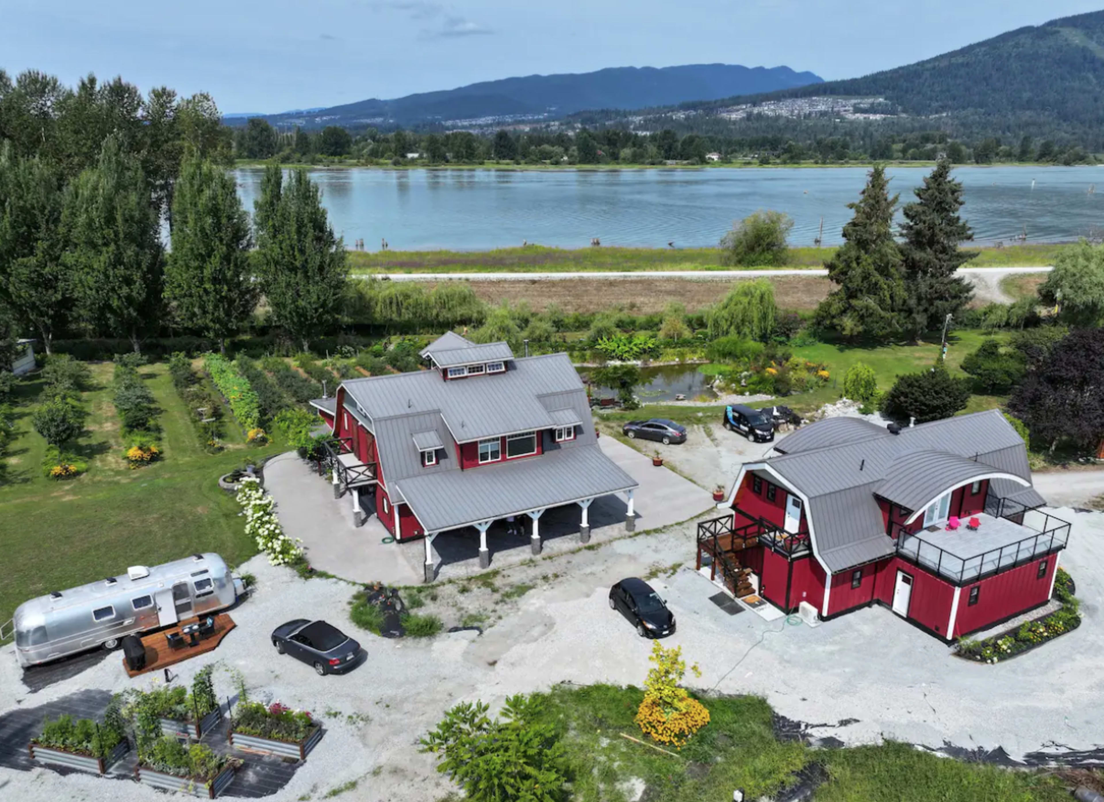 The image shows a red two-story house and a silver Airstream trailer near a river, surrounded by landscaped gardens and parked vehicles.
