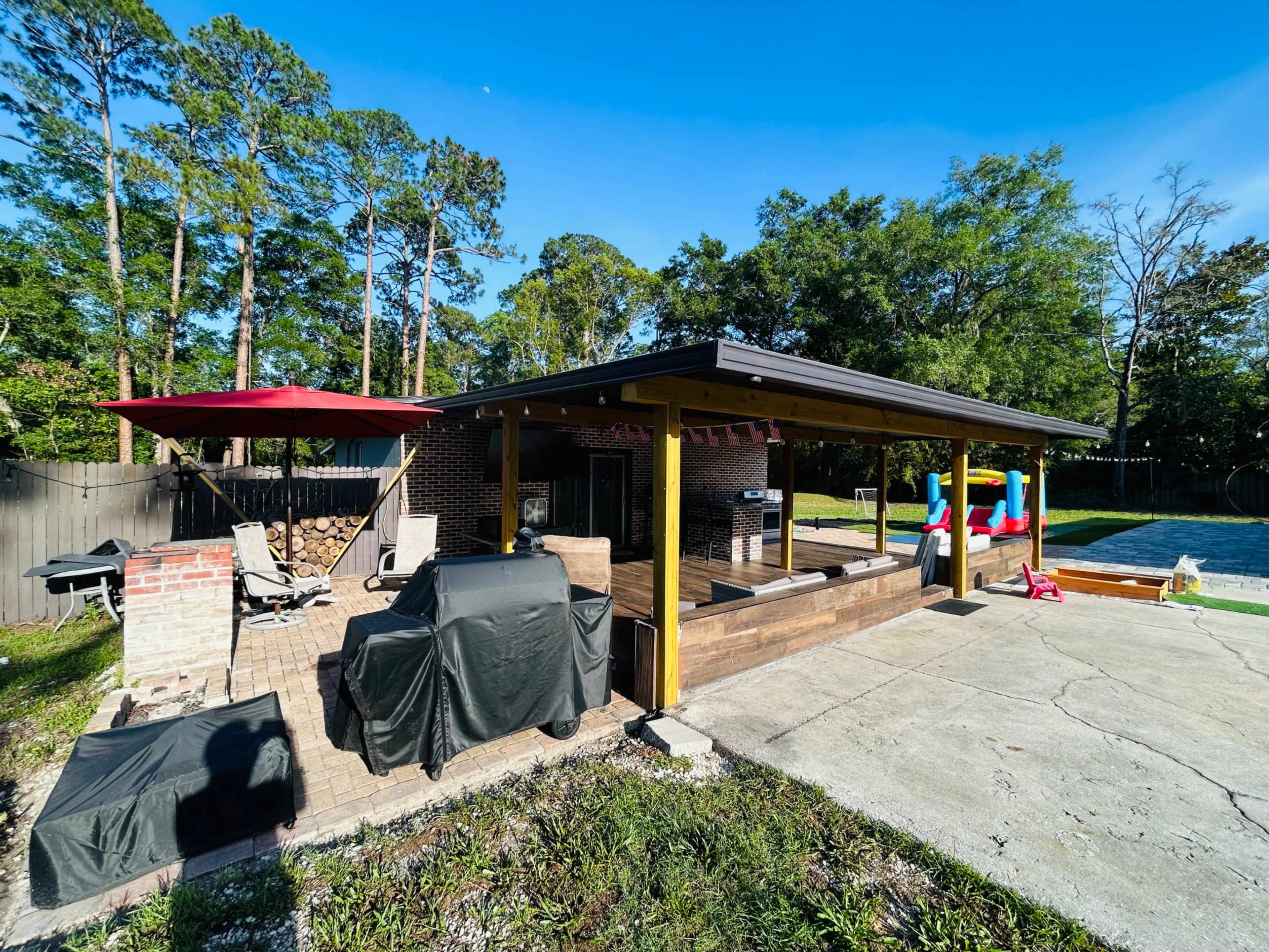 The image shows a backyard patio area with a grill, seating, and a pool visible in the background, surrounded by trees.