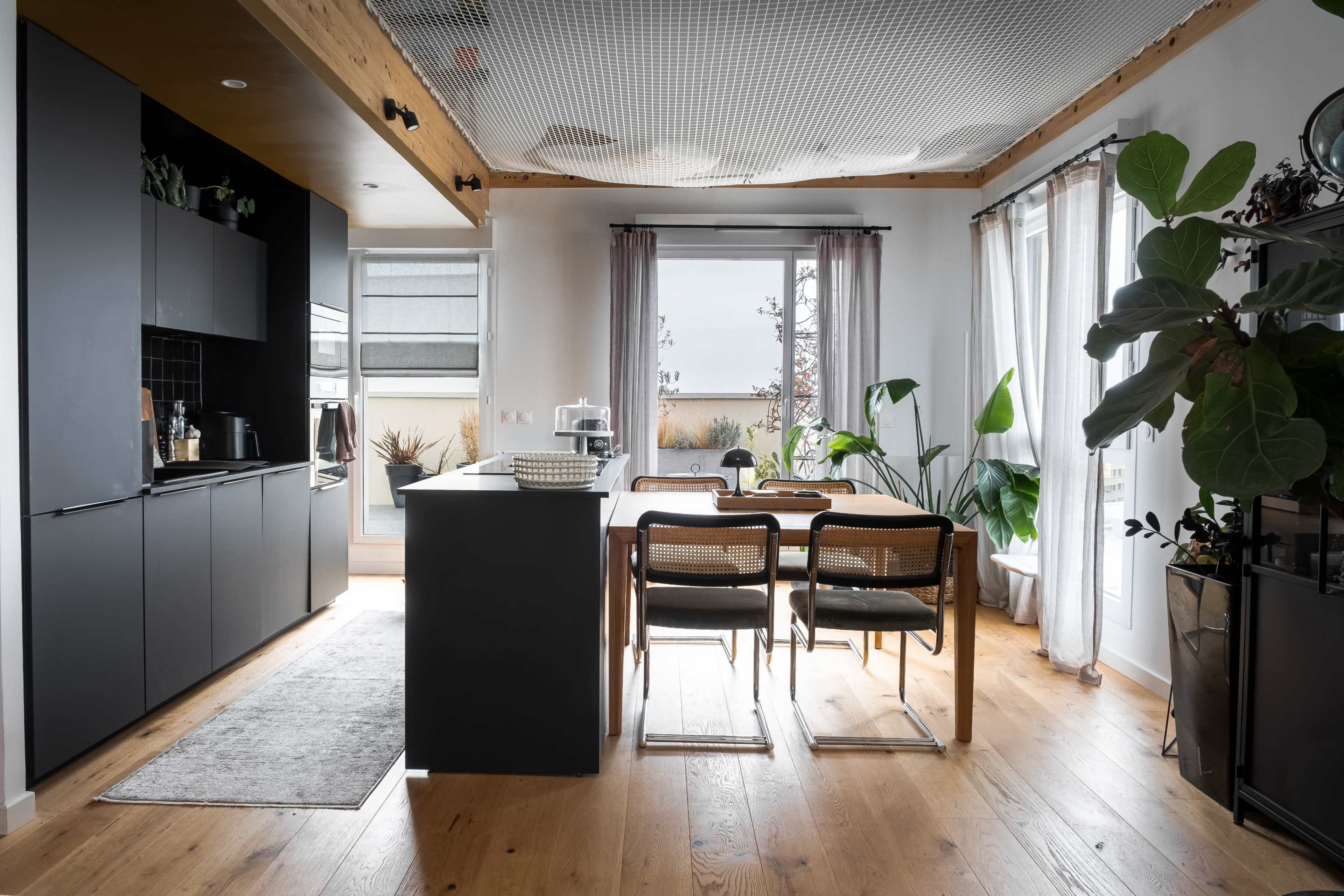 The image shows a modern kitchen with dark cabinetry, a central island, and an adjacent dining area filled with plants.