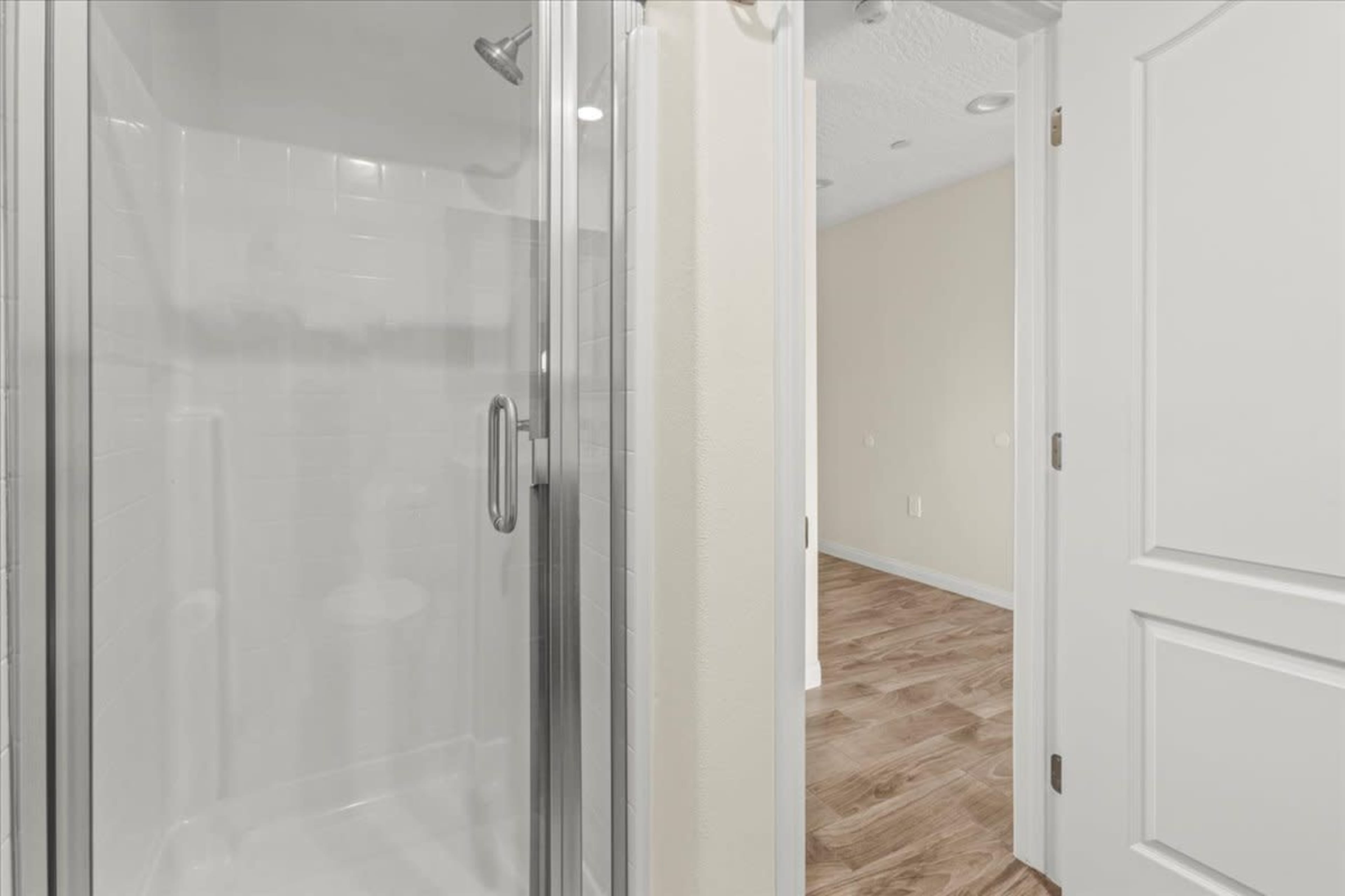 A clear glass shower enclosure adjacent to a partially visible room with a wooden floor and light-colored walls.