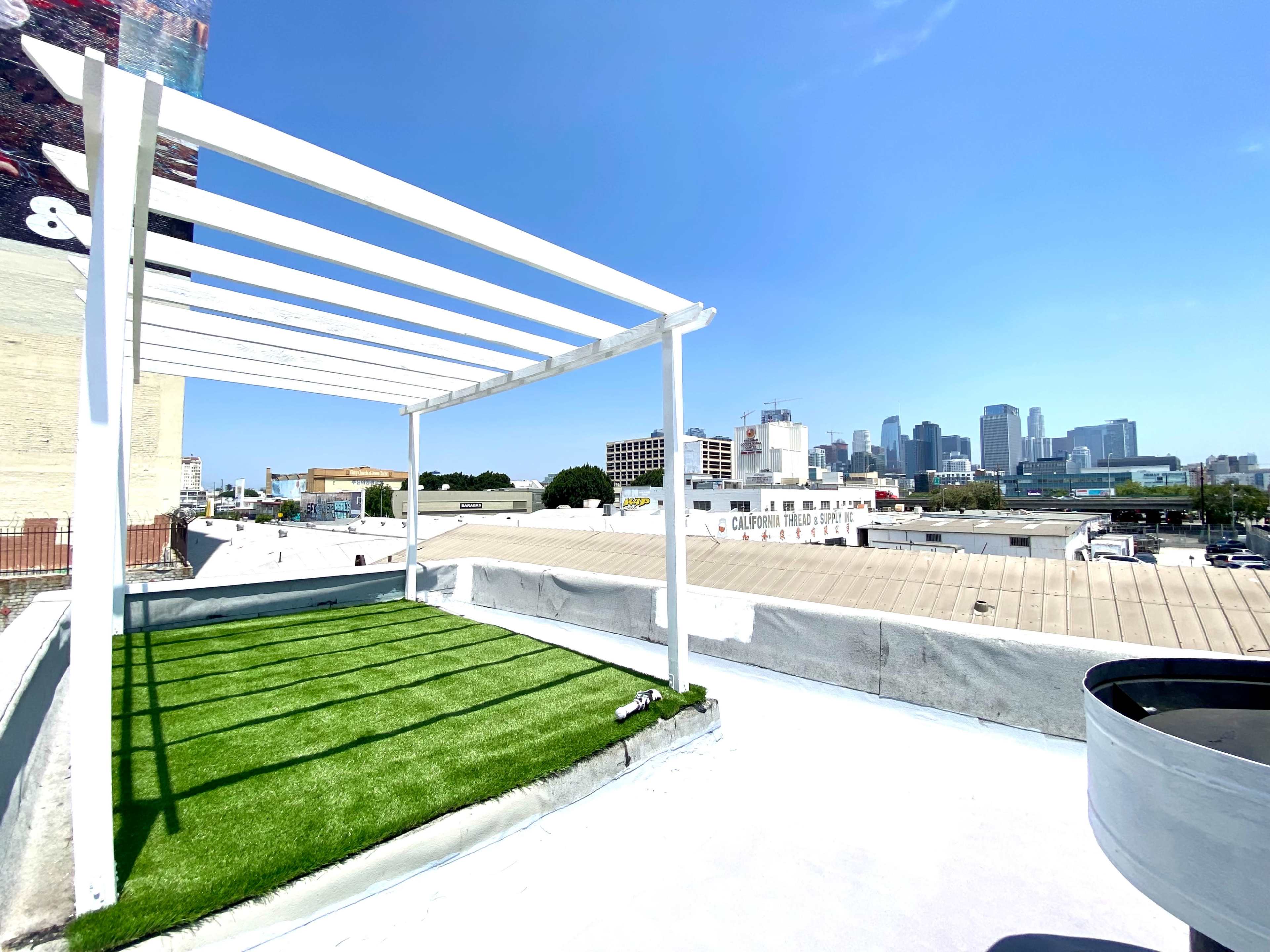 A rooftop terrace with artificial grass and a white pergola overlooks a city skyline under a clear blue sky.