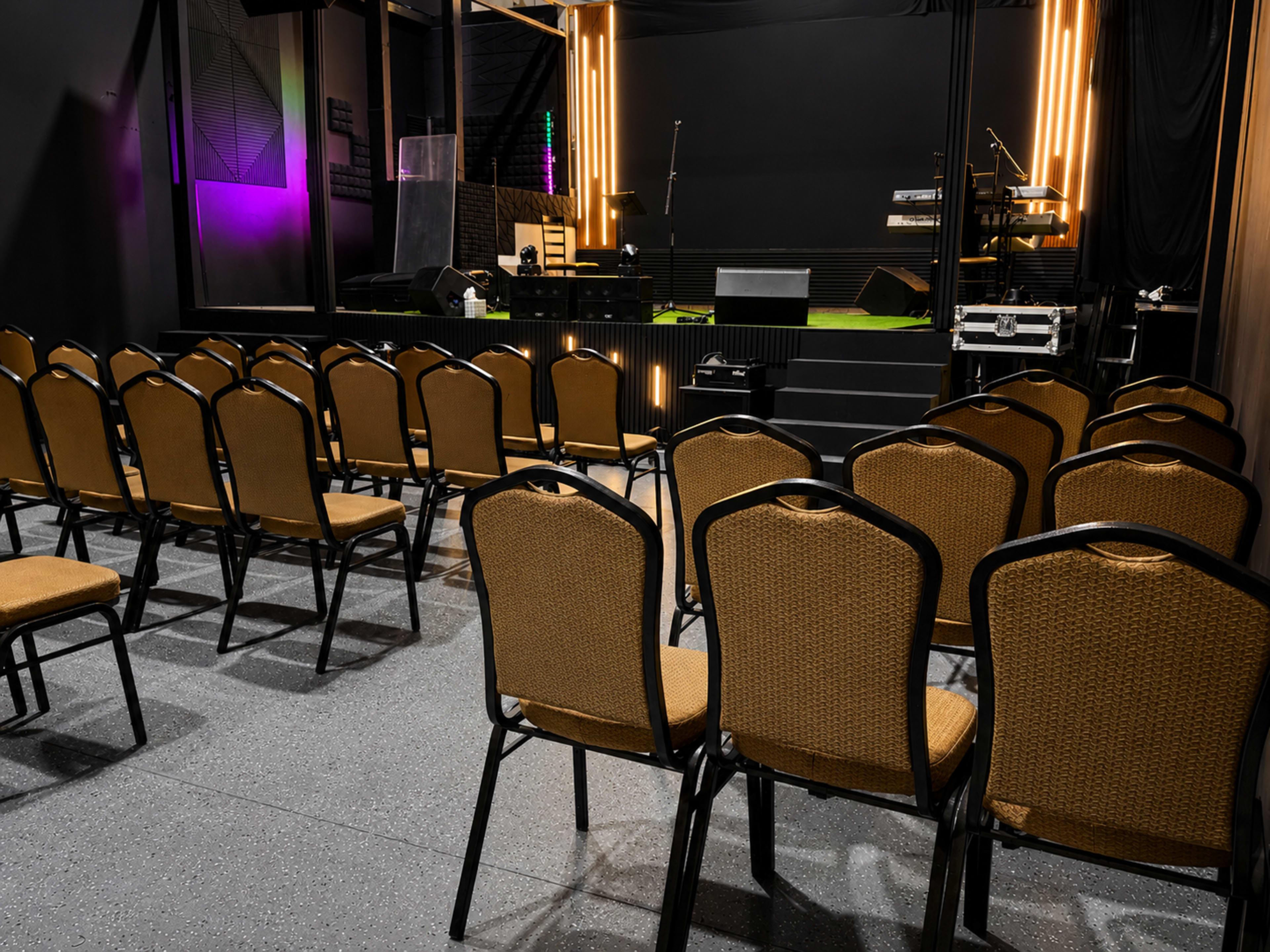 The image shows a row of empty chairs arranged facing a small stage with musical instruments in a dimly lit venue.