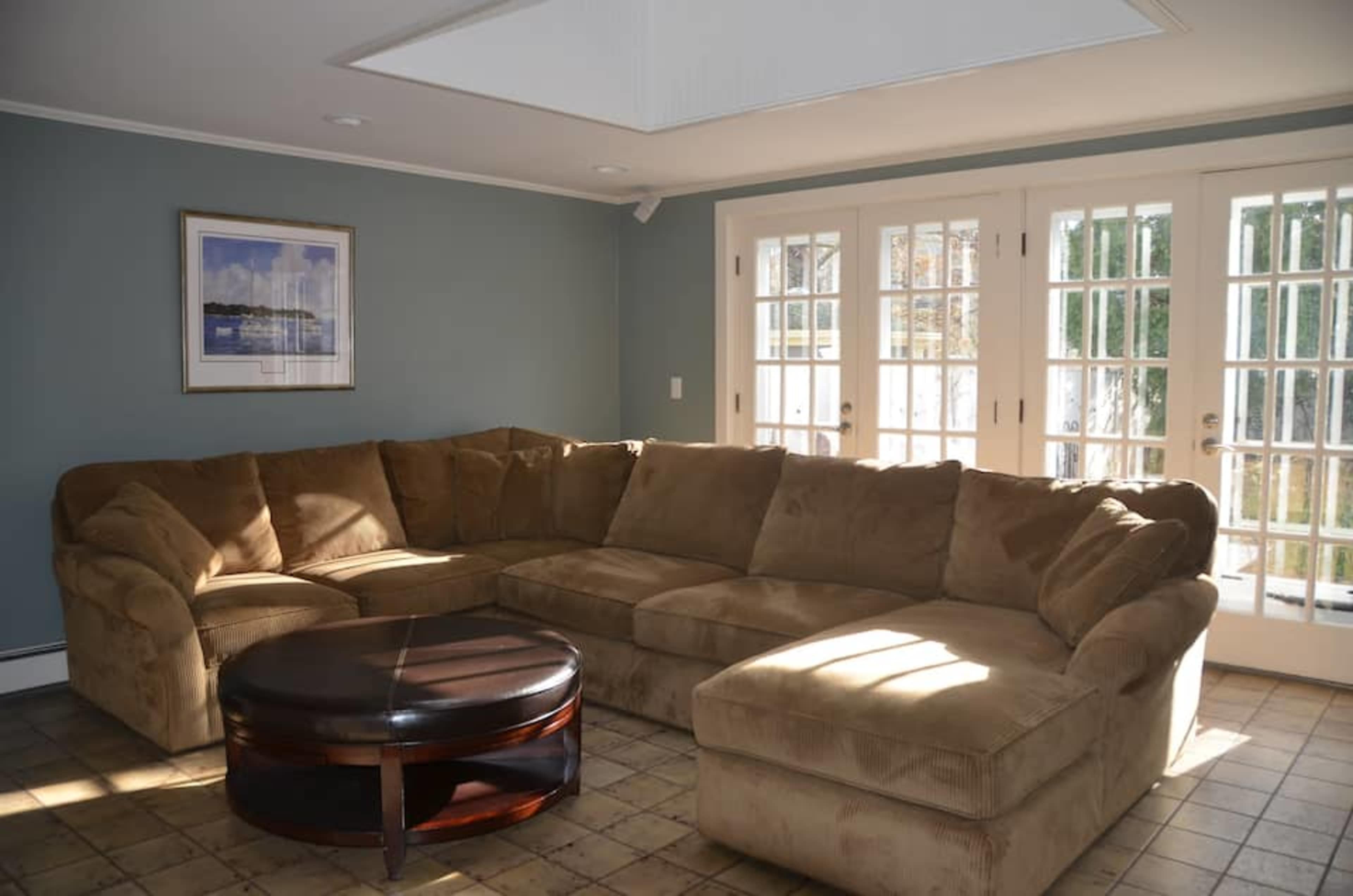 A spacious living room with a large, brown sectional sofa and a round coffee table, illuminated by natural light from double French doors.