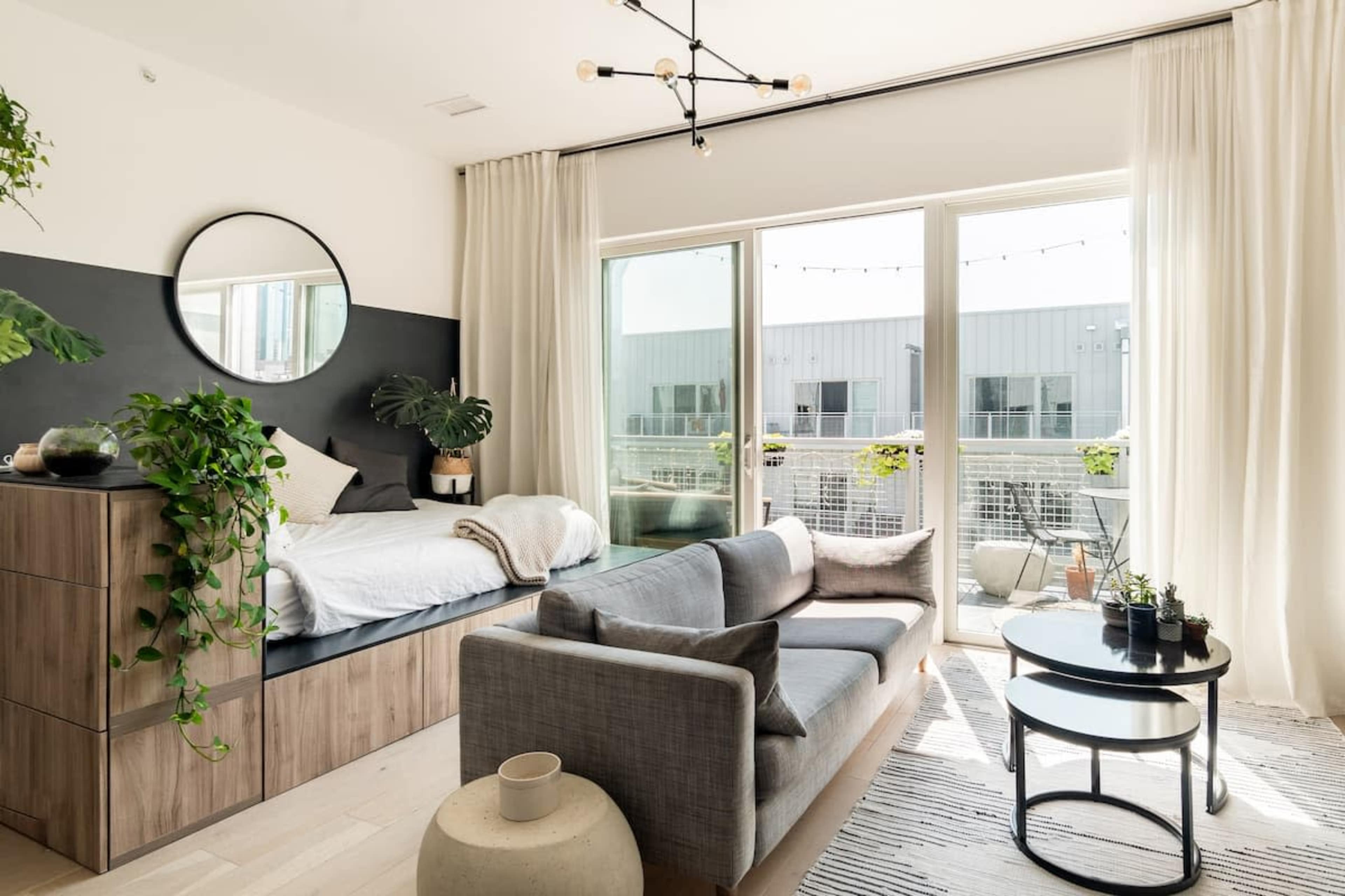 A modern bedroom with a bed, a sofa, and large glass doors leading to a balcony, adorned with greenery and bright natural light.