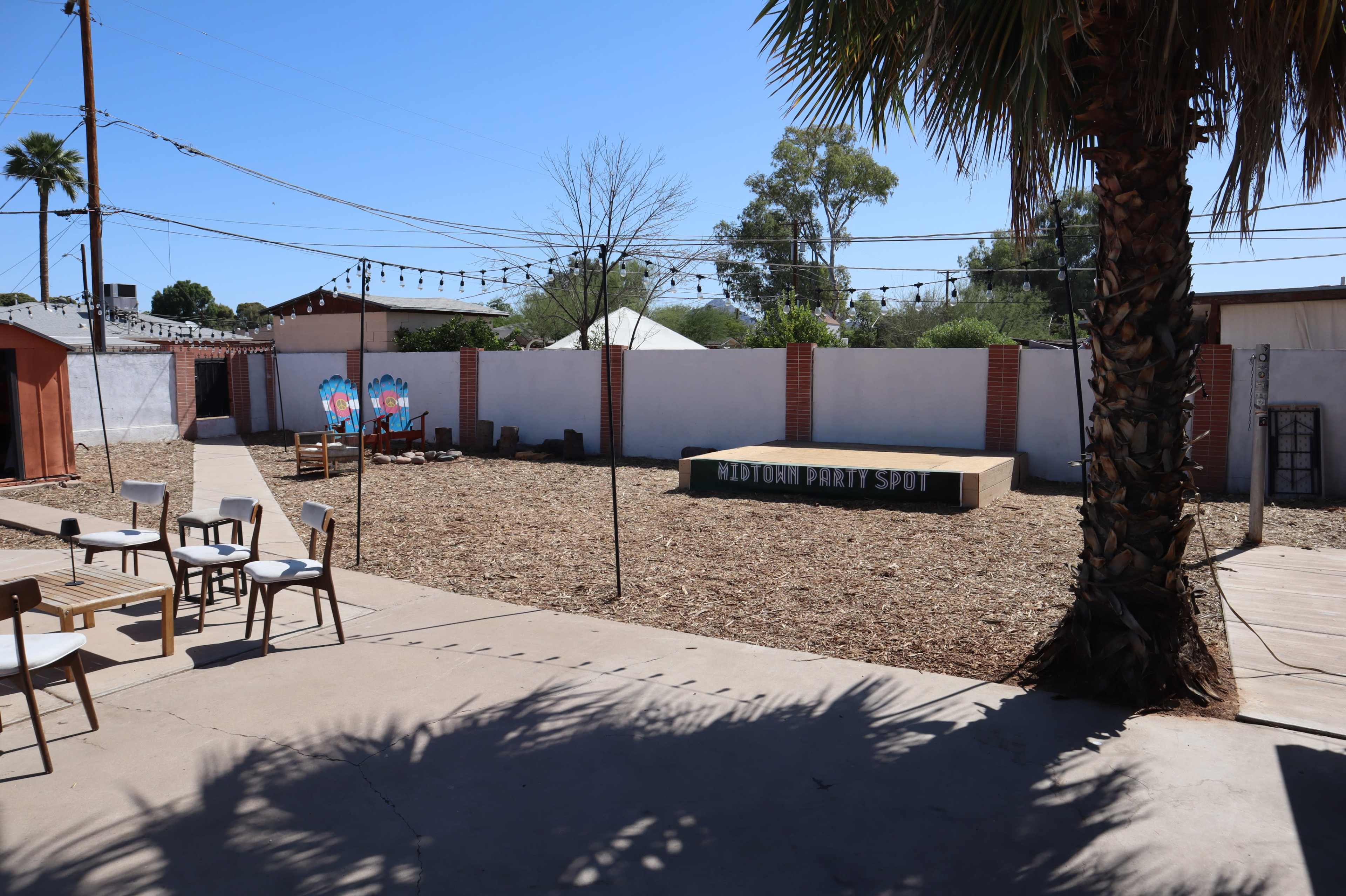 The image shows an outdoor space with a wooden performance stage, surrounded by a gravel area, chairs set up in the foreground, and a wall featuring colorful murals.