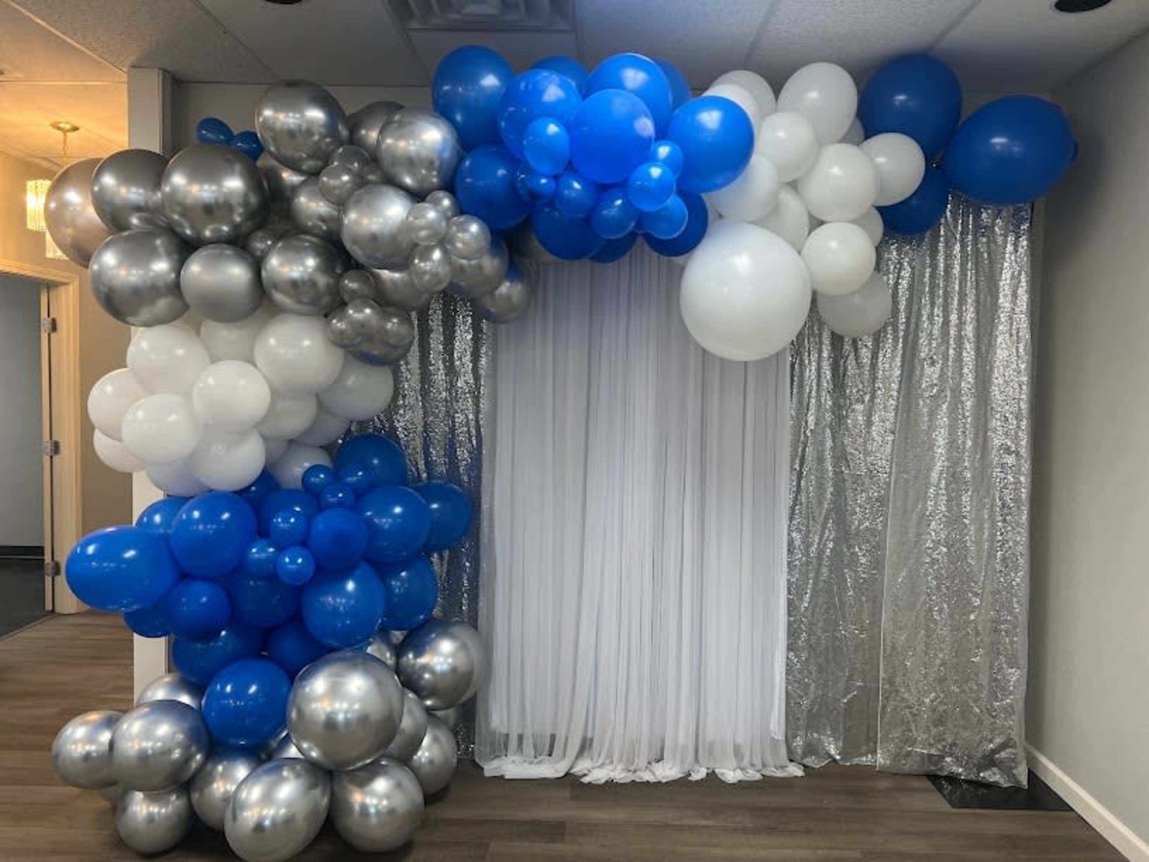 A decorative archway made of blue, white, and silver balloons, set against a backdrop of white drapery and a silver curtain.
