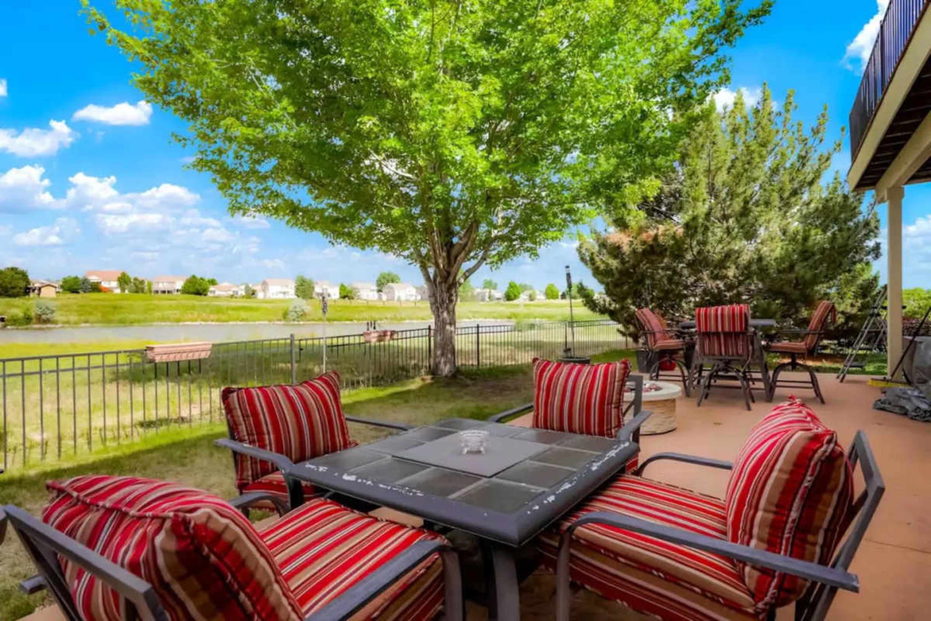 A patio features a table surrounded by chairs, overlooking a pond and a grassy area with trees in the background.
