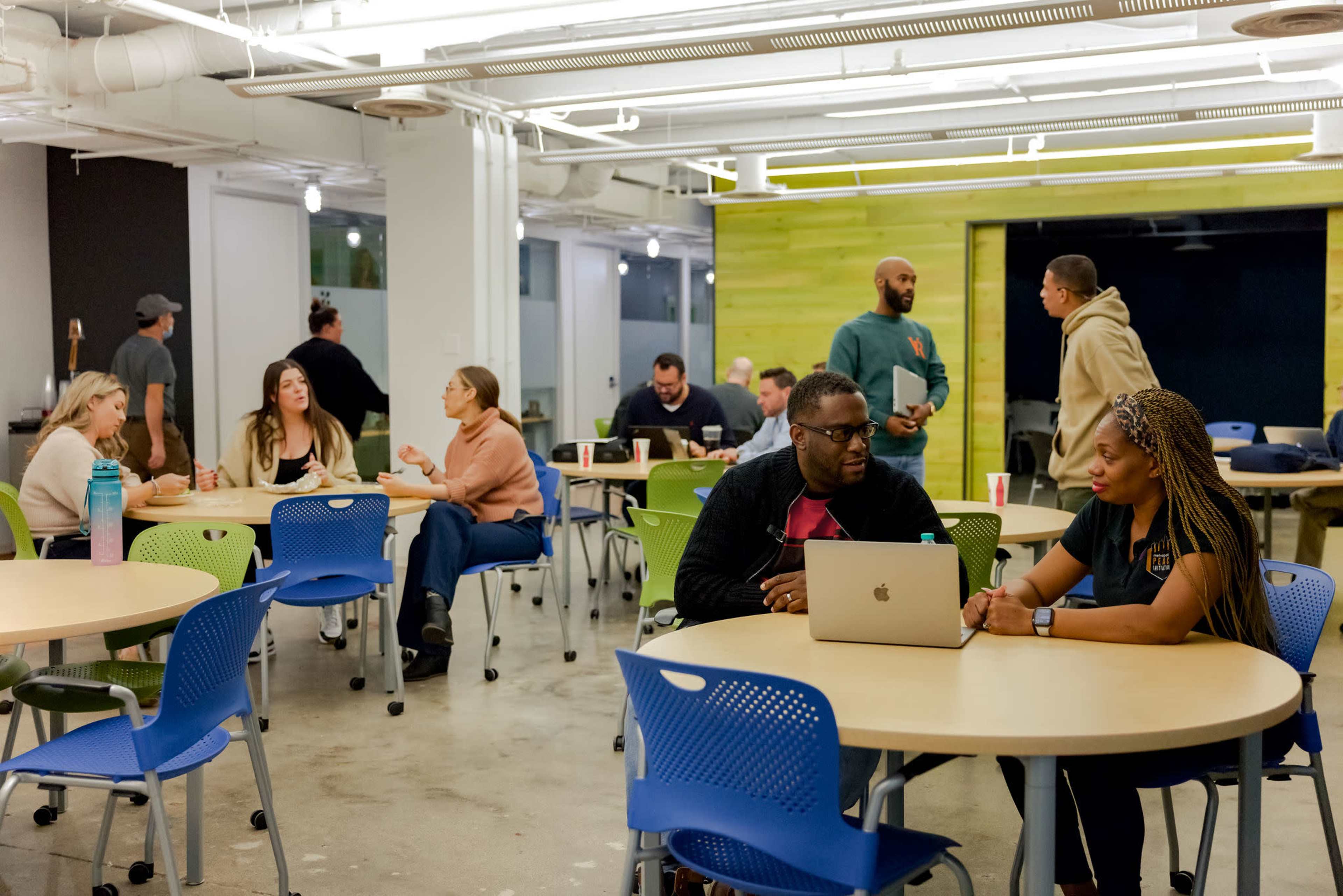 A group of people is engaged in conversations and work at tables in a modern, brightly lit workspace.