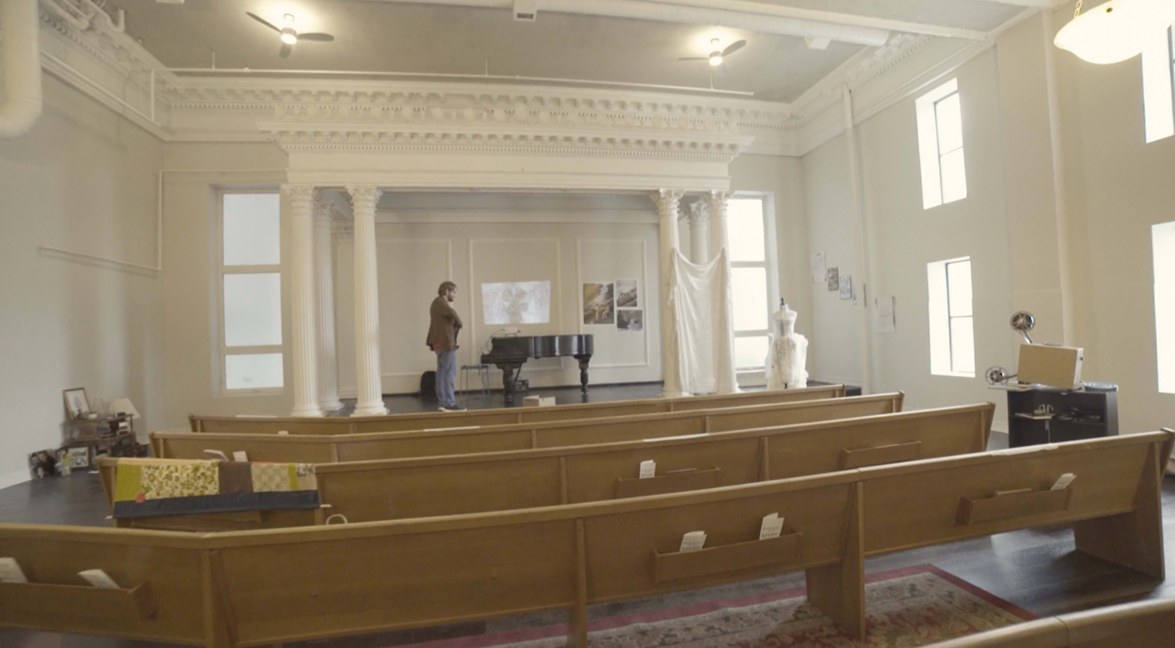 A person stands next to a grand piano on a raised stage surrounded by wooden pews in a spacious room with large windows and high ceilings.