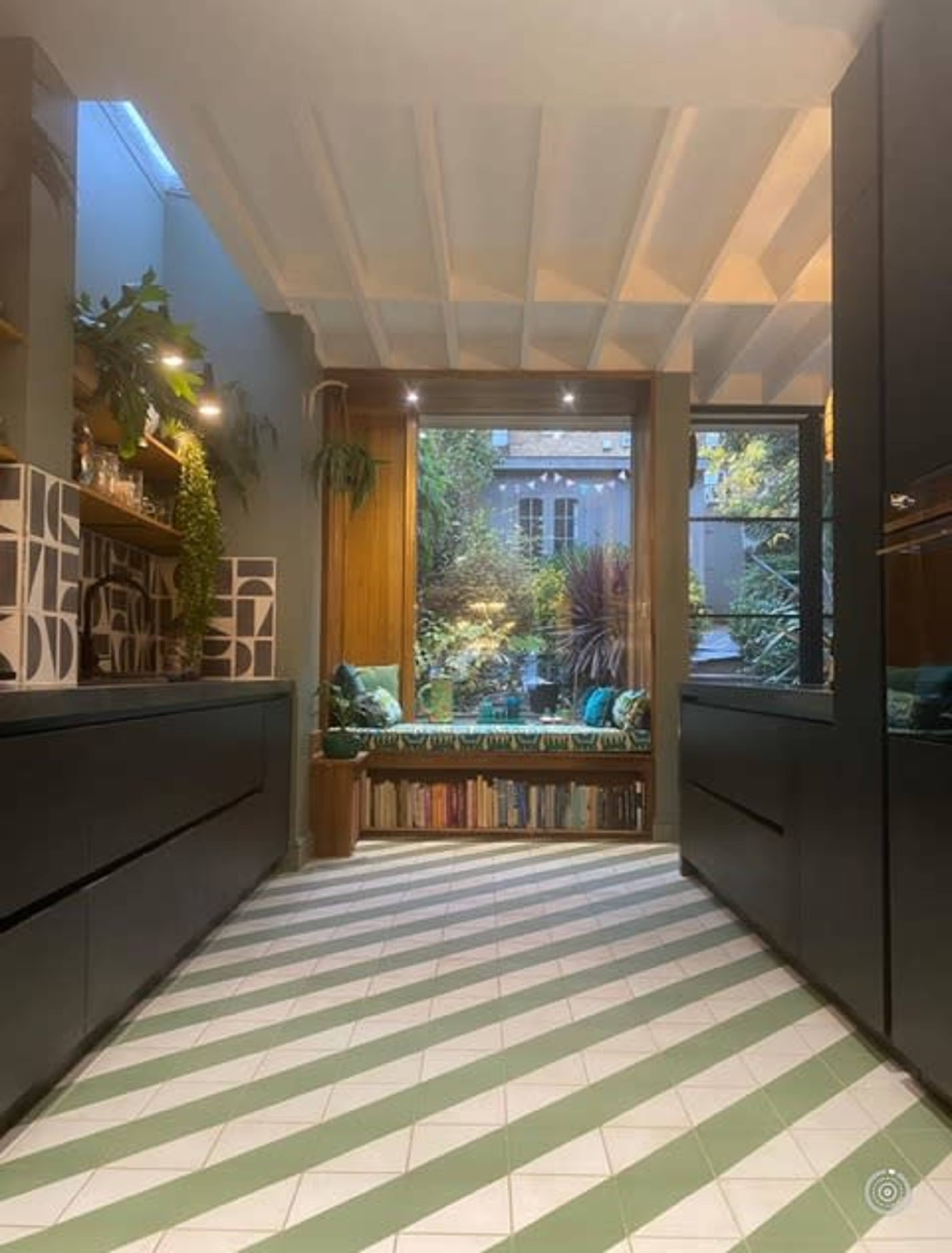 A modern kitchen with a patterned green and white floor, leading to a window seating area that overlooks a garden.