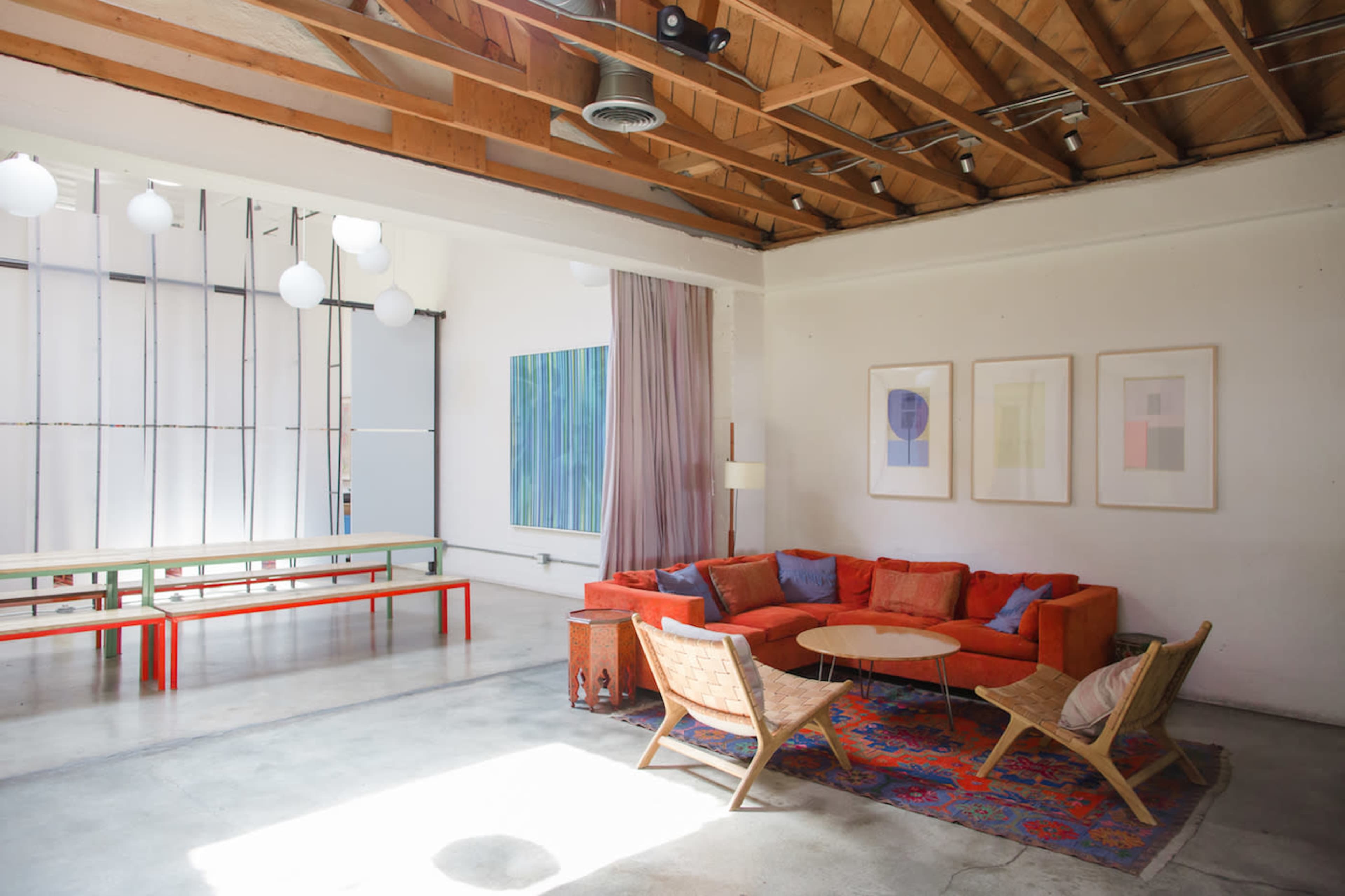 A bright, open living space featuring an orange sectional sofa, a round coffee table, and wooden chairs, with large windows and minimalist decor.