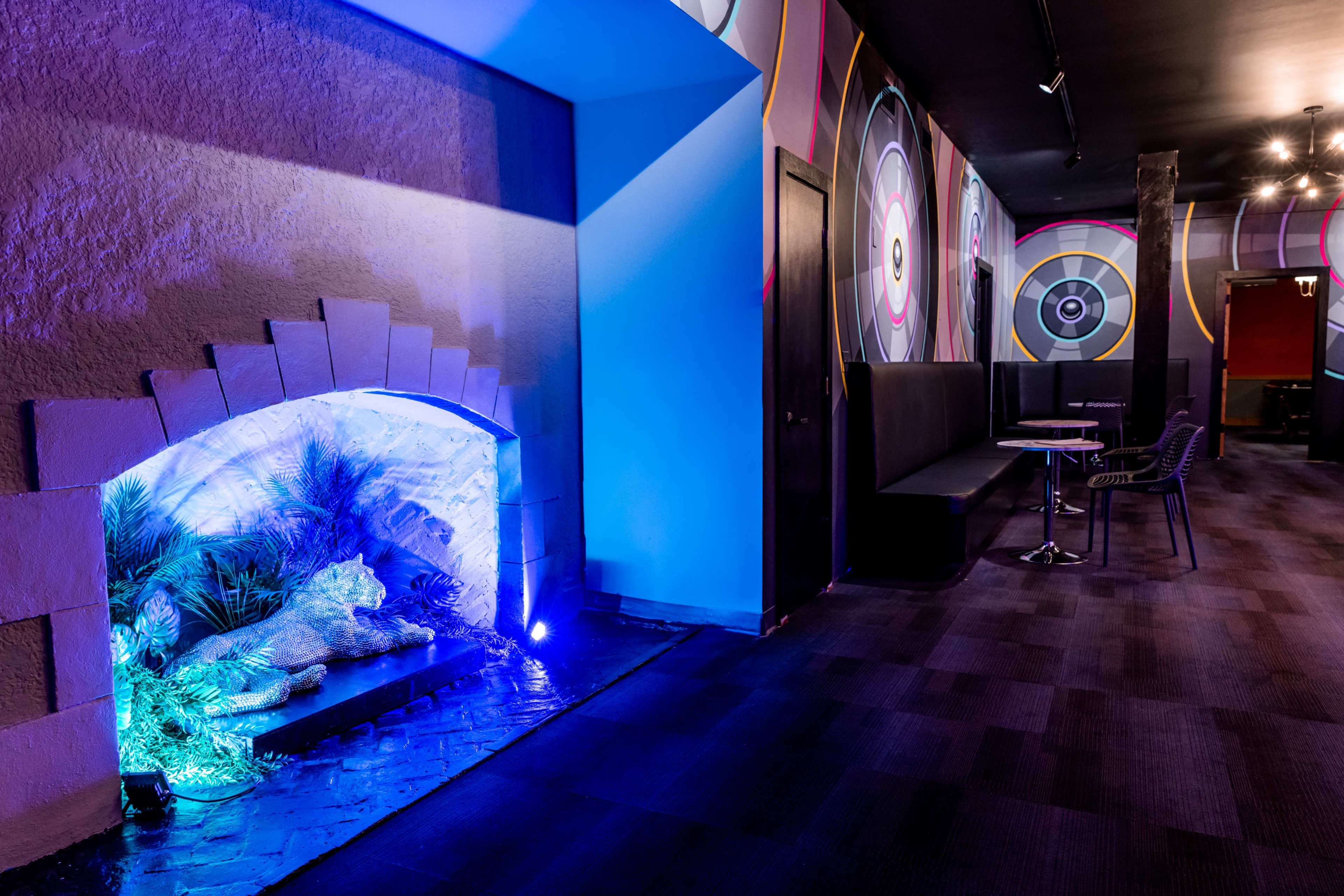 The image features a brightly lit hallway with colorful wall designs, a seating area, and a decorative lion statue surrounded by plants illuminated by blue lighting.
