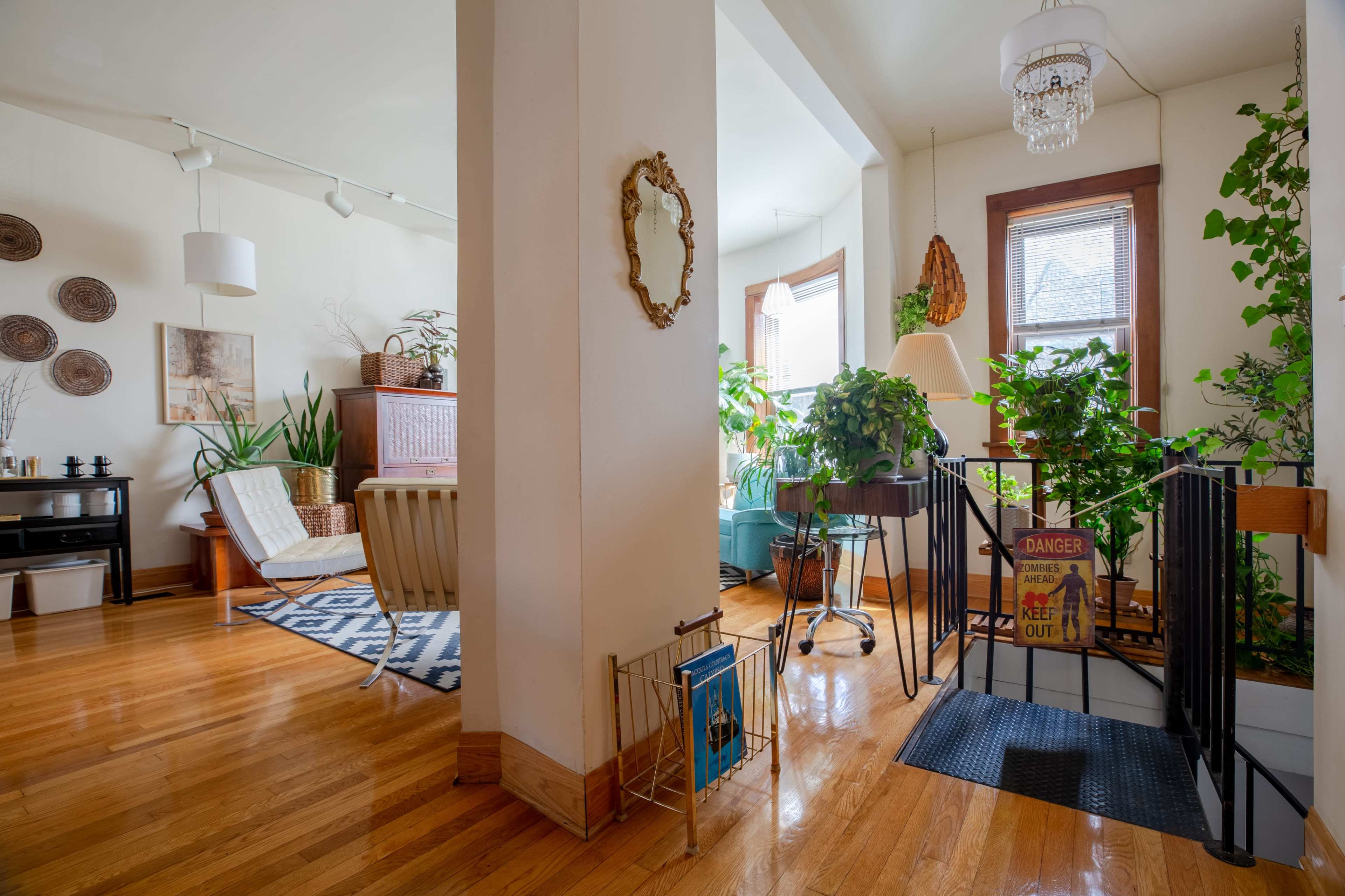 The image shows a cozy living space filled with plants, wooden floors, and a mix of furniture, including a chair, a lamp, and a decorative mirror.