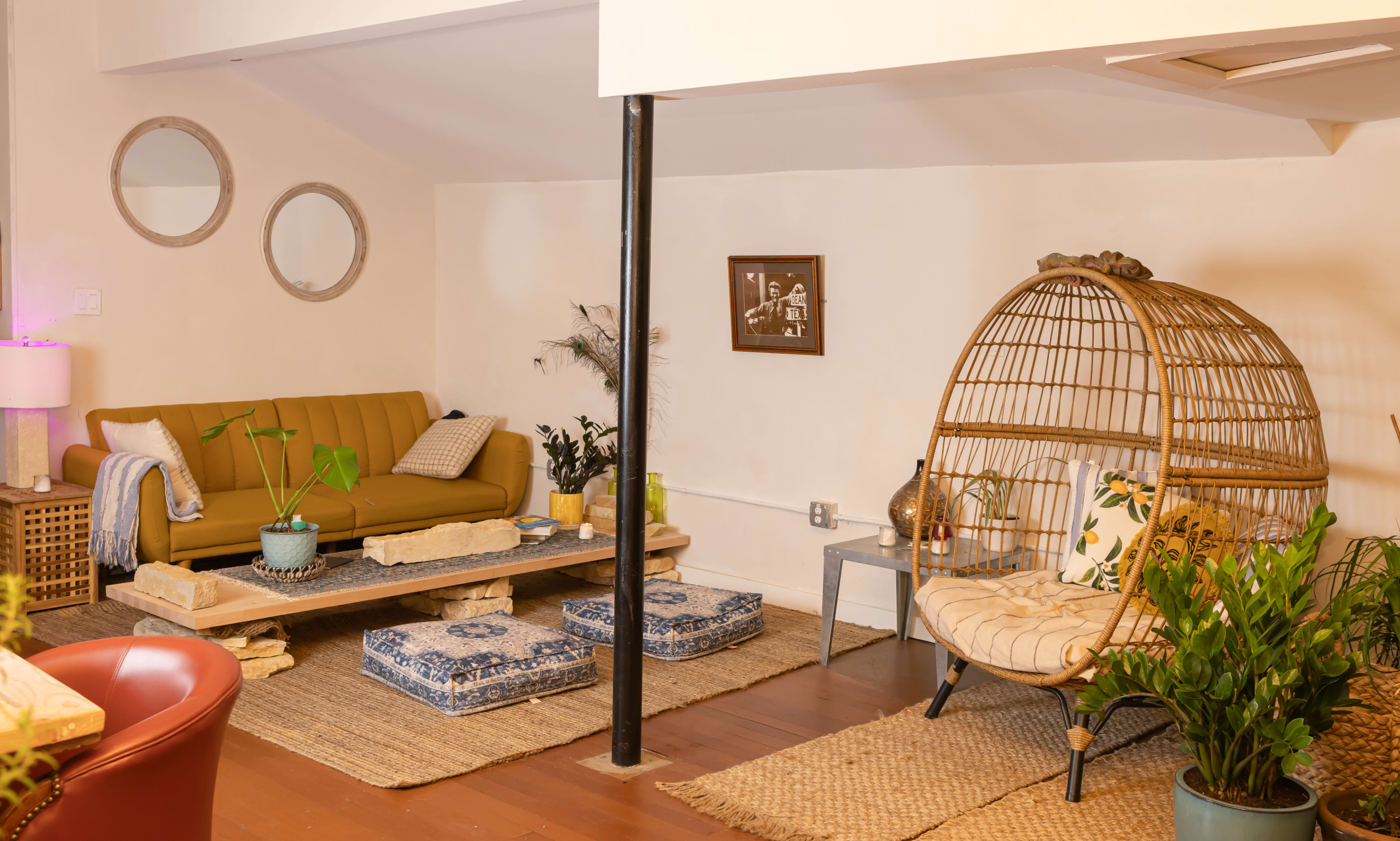 The image shows a cozy living area with a yellow couch, a circular rattan chair, stone coffee tables, and various plants.
