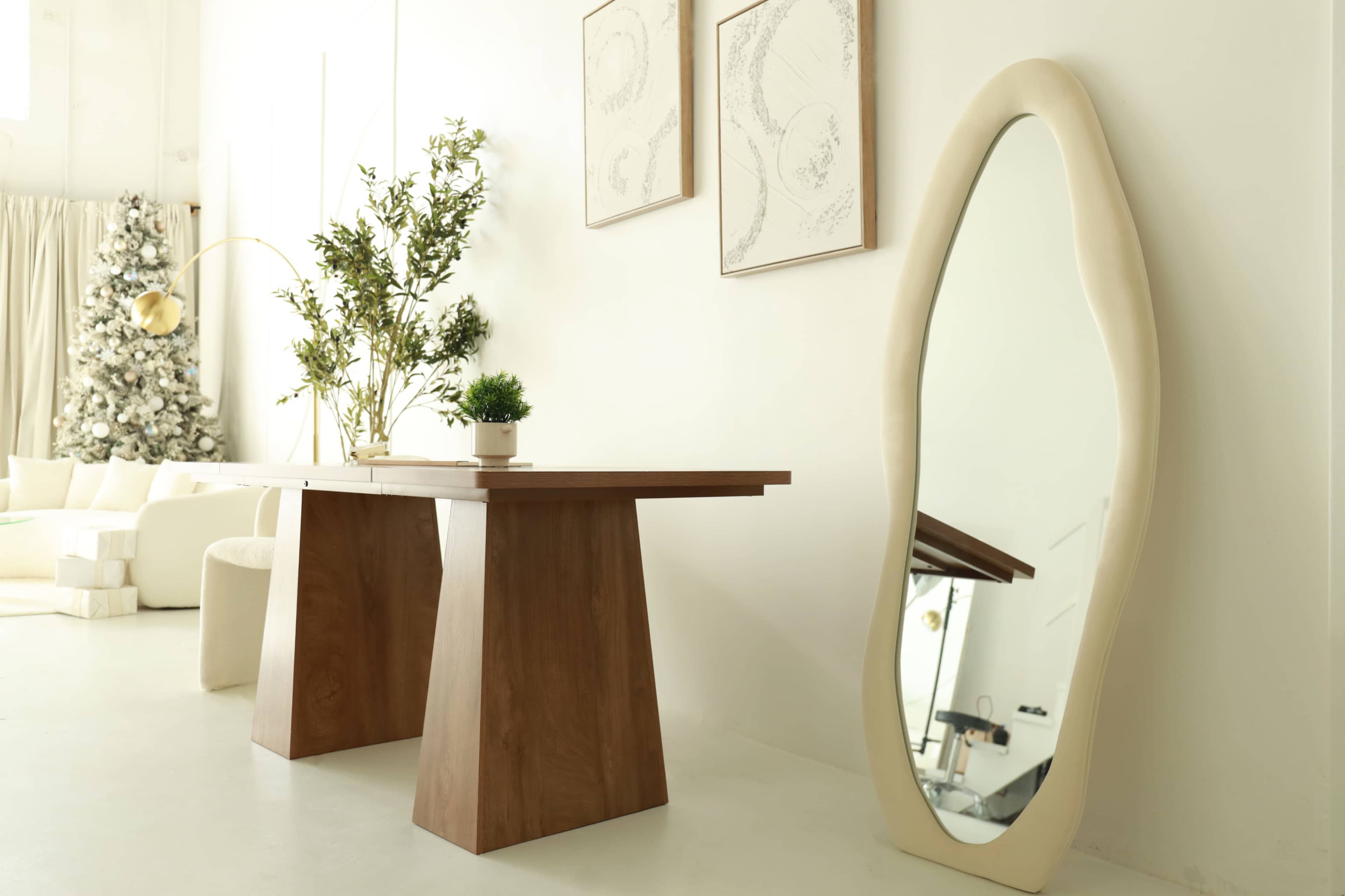 A wooden table with a geometric design is positioned next to an abstractly shaped mirror against a white wall, complemented by decorative artwork and a plant.