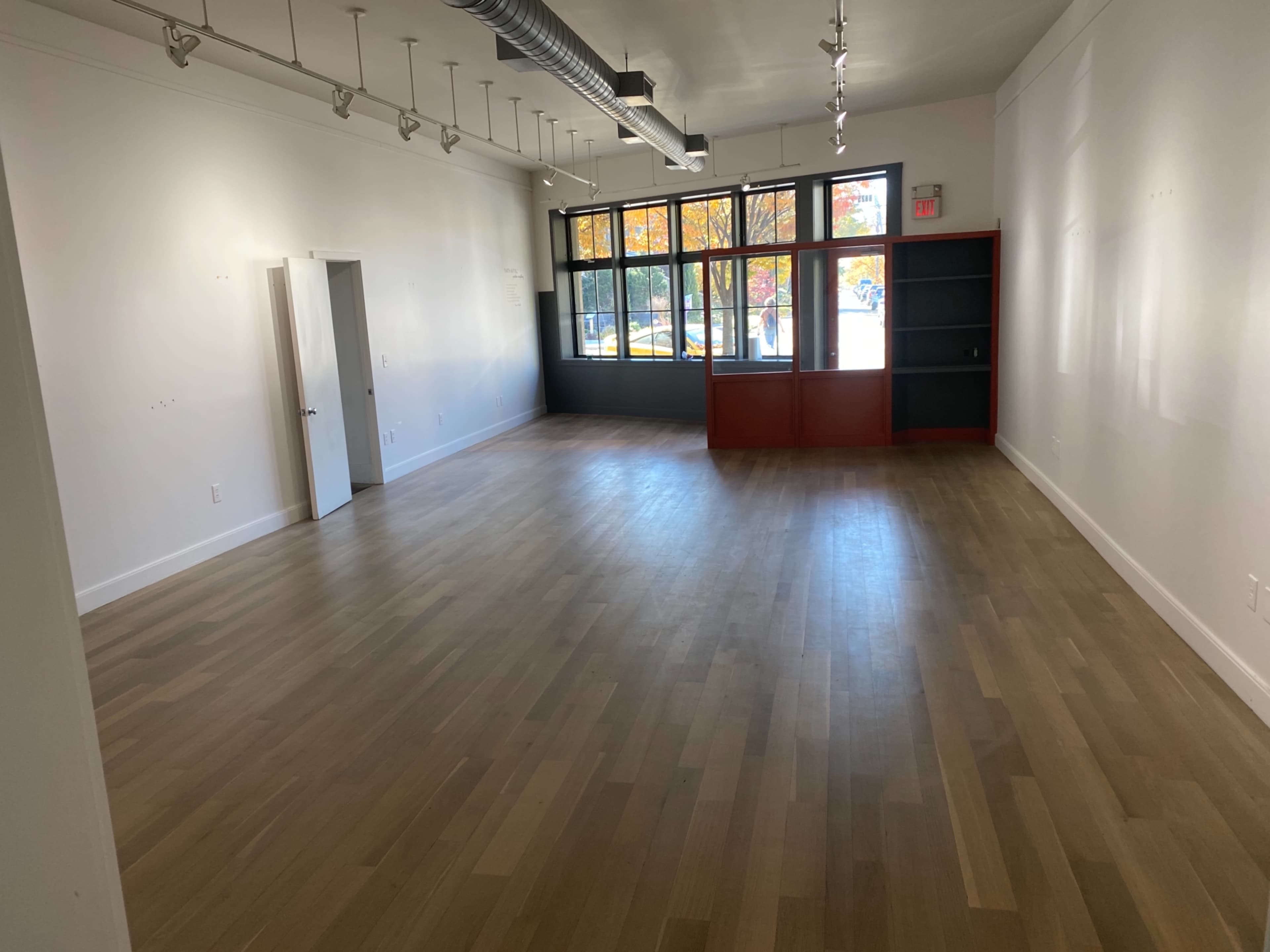 An empty room with wooden flooring, large windows, and a red bookshelf against one wall.