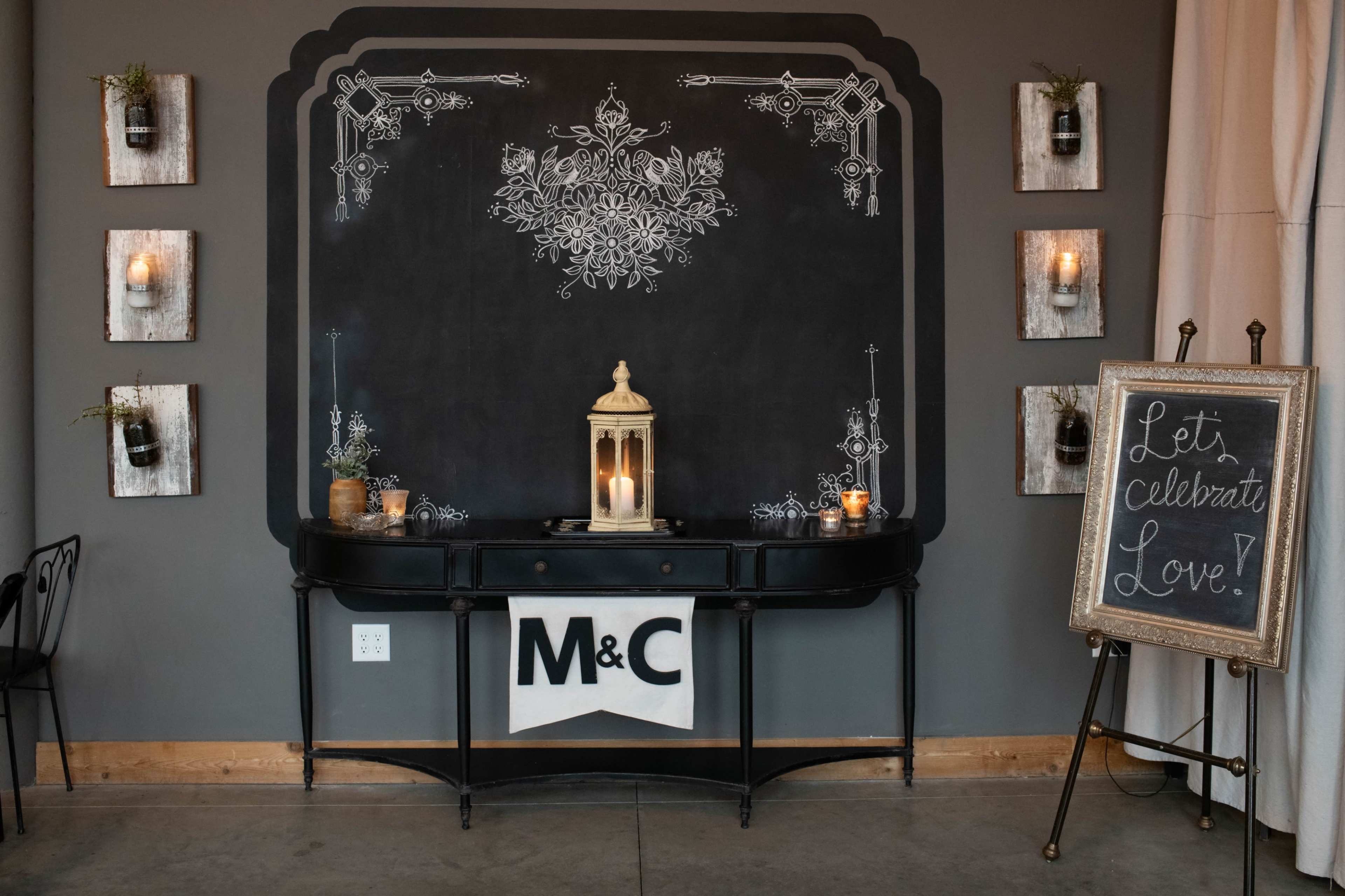 A decorative chalkboard wall displays floral designs, flanked by candle-lit wall planters, with a table beneath featuring a lantern and a sign that invites celebration.