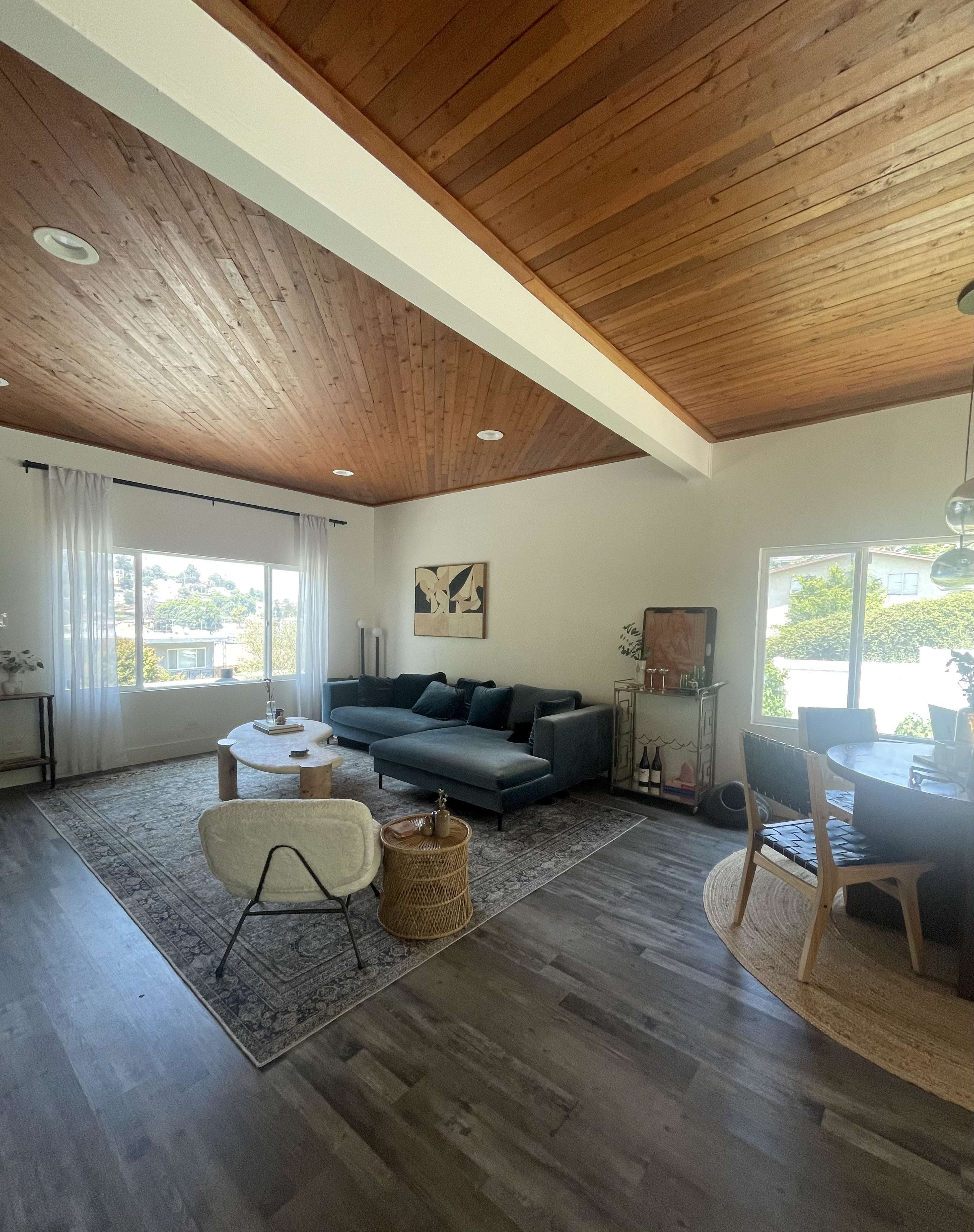 A bright, contemporary living area features a dark sectional sofa, a round coffee table, and a dining area with a wooden table, all set against a backdrop of wooden ceilings and large windows.